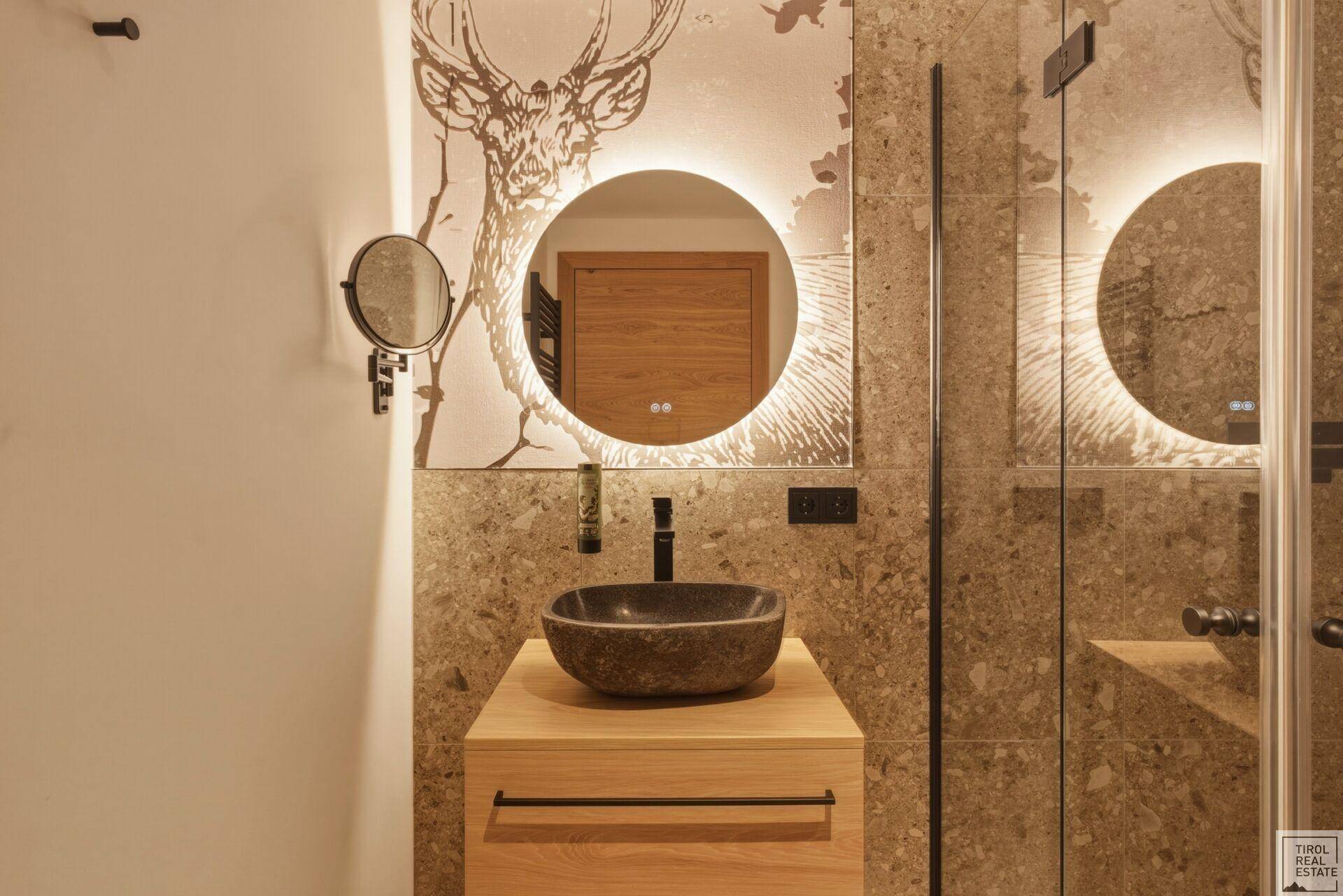 Detailansicht des Badezimmers mit Steinwaschbecken, beleuchtetem Spiegel und Hirschmotiv an der Wand.