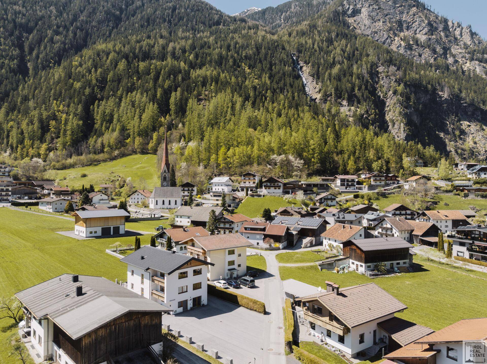 Malerische Luftaufnahme eines alpinen Dorfes, umgeben von grünen Wiesen und majestätischen Bergen.