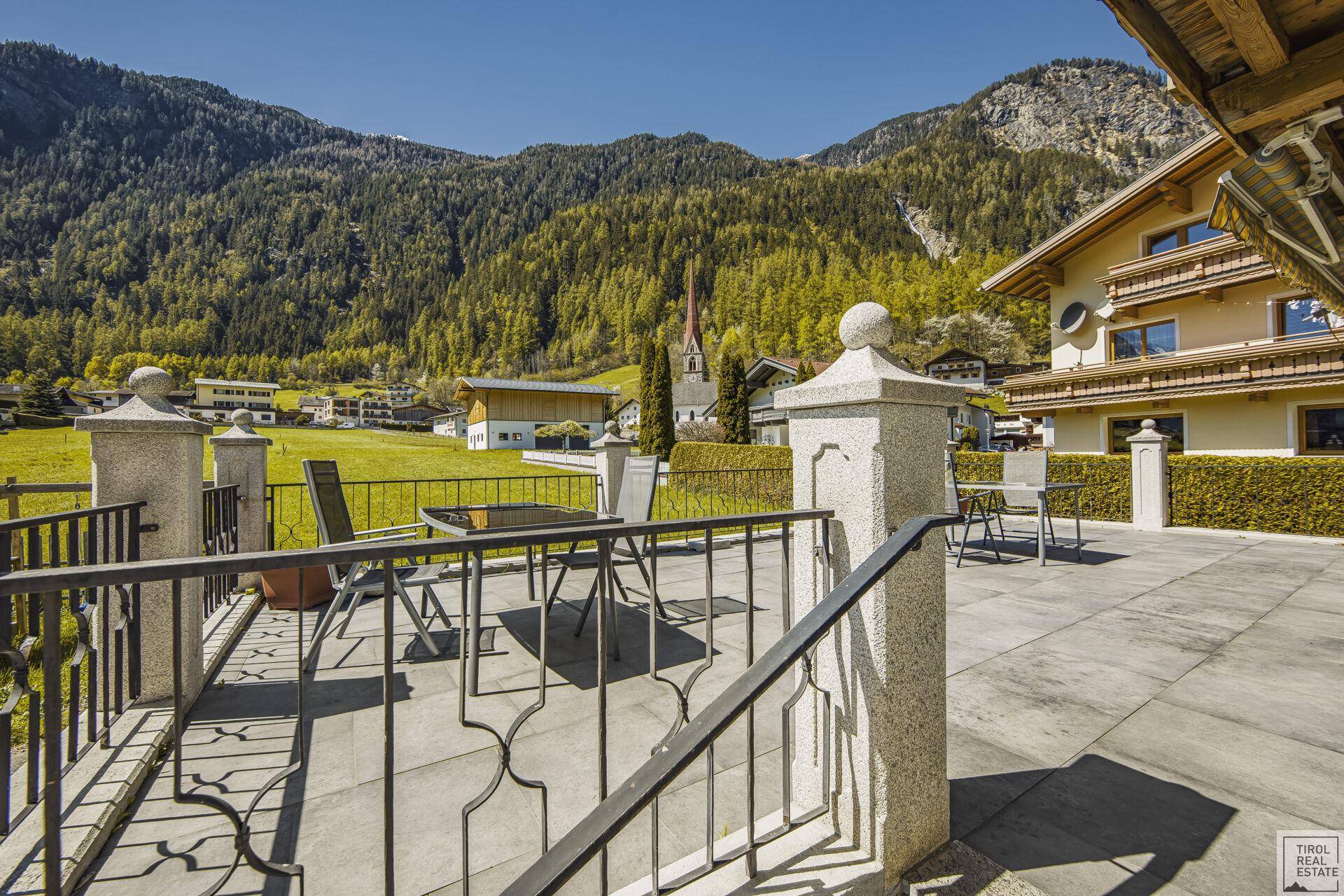 Große gepflasterte Terrasse mit Sitzgelegenheiten und Blick auf die Dorfkirche und Berge.