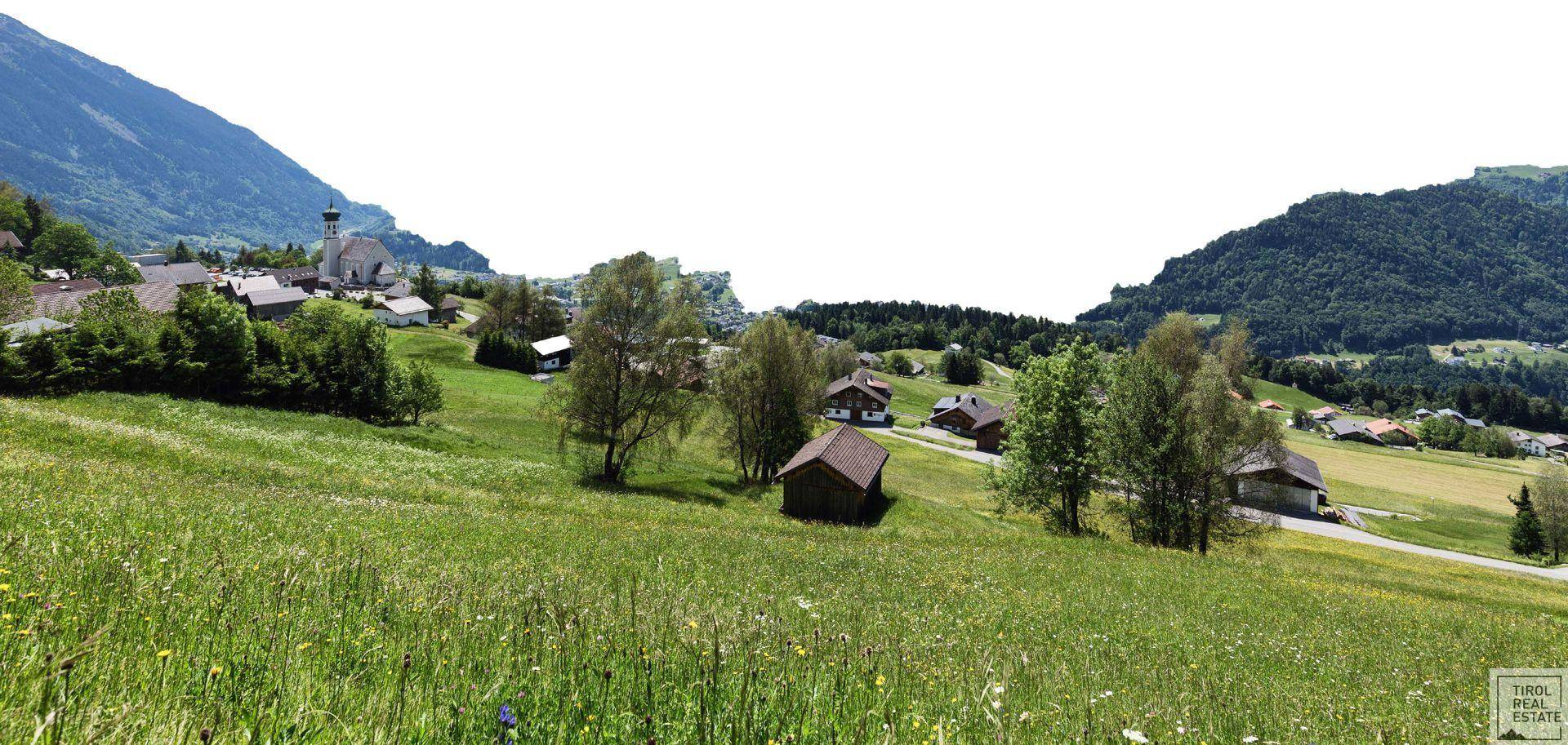 Weitläufige grüne Landschaft mit verstreuten Häusern und Bergen im Hintergrund.