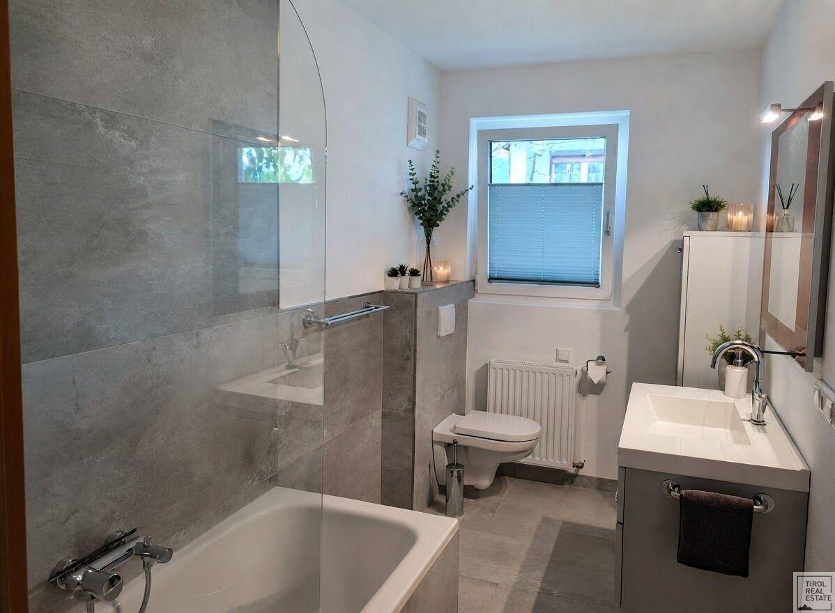 Helles Badezimmer mit Badewanne, Toilette, modernem Waschtisch und grauen Fliesen, gut beleuchtet.