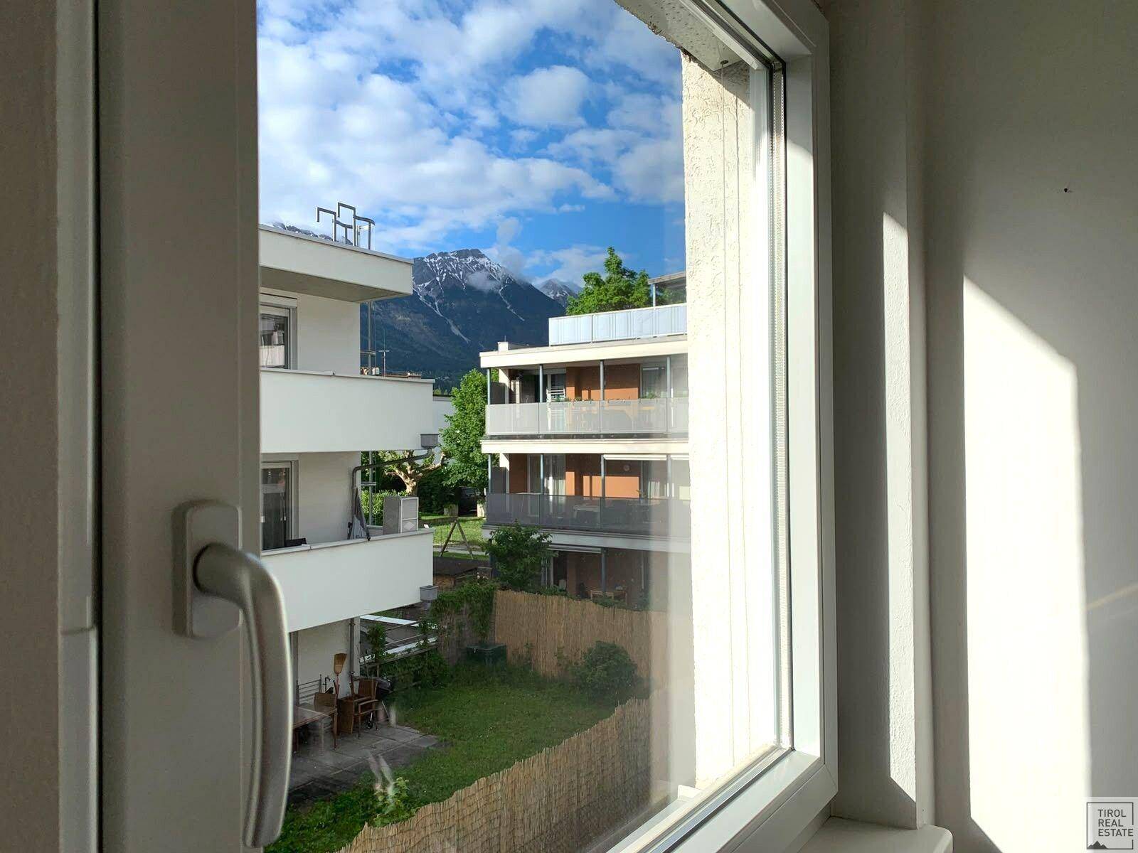 Panoramablick aus dem Fenster auf umliegende Gebäude, grüne Gärten und schneebedeckte Berge.