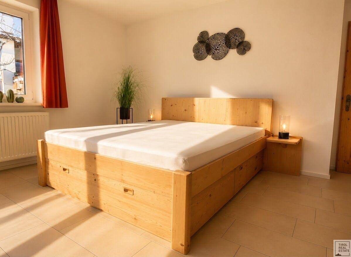 Gemütliches Schlafzimmer mit hellem Holzbettrahmen, Fliesenboden und einem Fenster, das viel Licht hereinlässt.