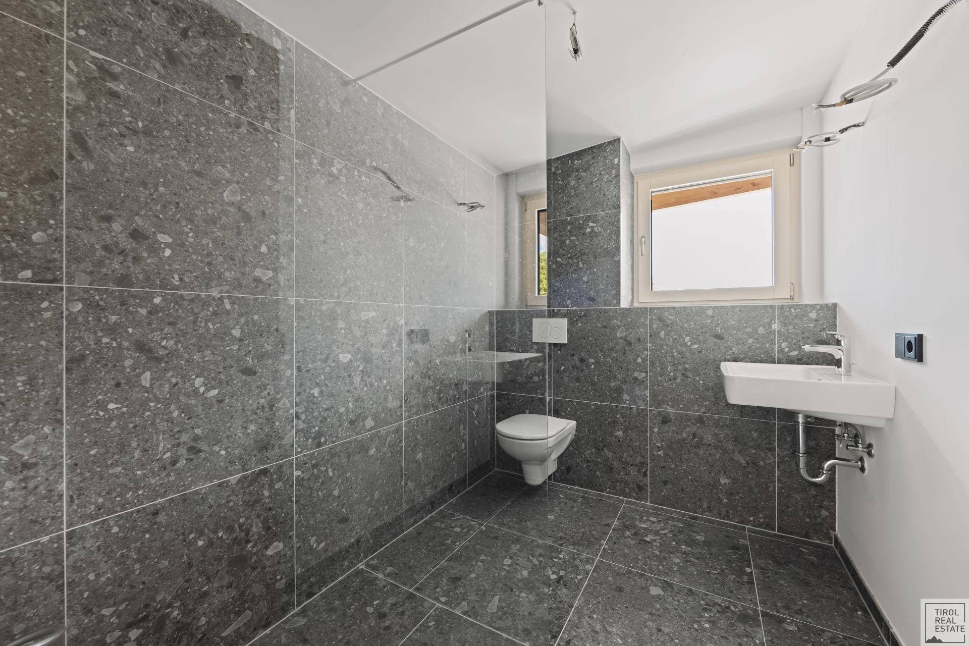 Geräumiges Badezimmer mit grauen Fliesen, Dusche, WC und Fenster für Tageslicht.