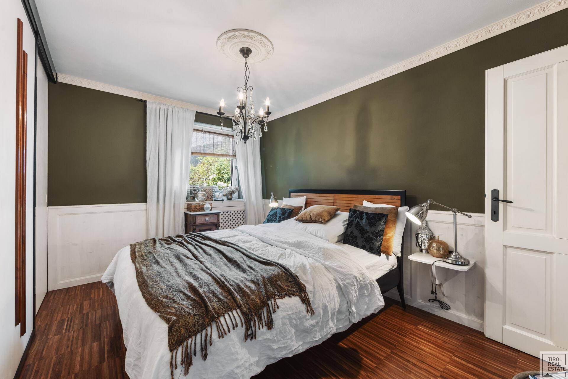Gemütliches Schlafzimmer mit dunkelgrünen Wänden, Holzboden und Fenster mit Blick ins Freie.