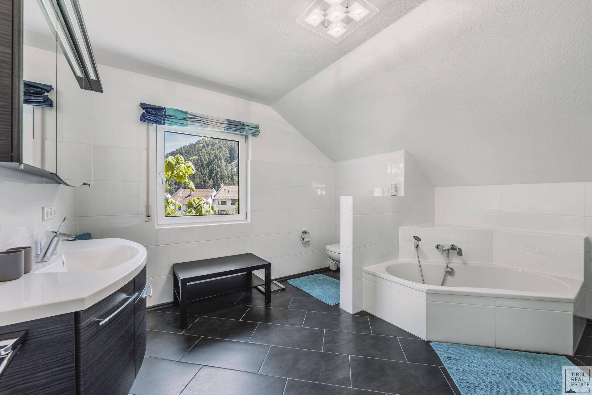 Modernes Badezimmer mit Eckbadewanne, dunklen Fliesen und Fenster mit Bergblick.