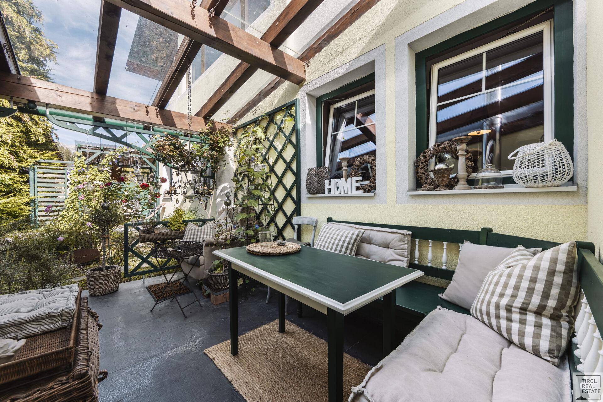 Überdachte Terrasse mit Glasdach, gemütlicher Sitzecke und üppiger grüner Bepflanzung.