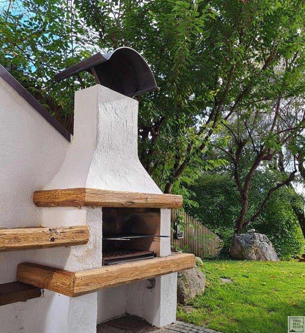 Gemütlicher Grillplatz im Garten mit gemauertem Grill und Holzelementen, umgeben von viel Grün.