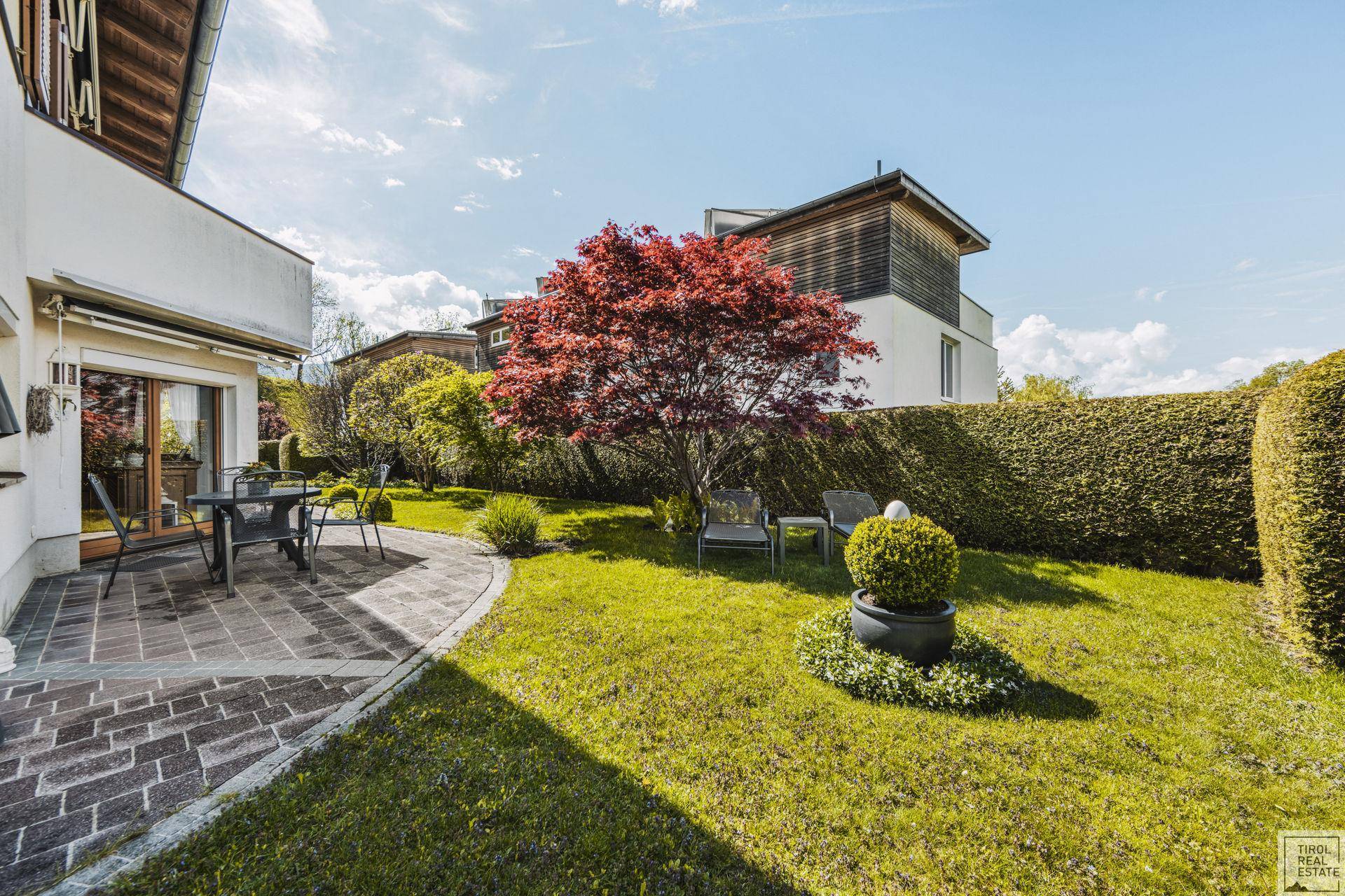 Gepflegter Garten mit rotem Baum, Rasenfläche und angrenzender gepflasterter Terrasse.