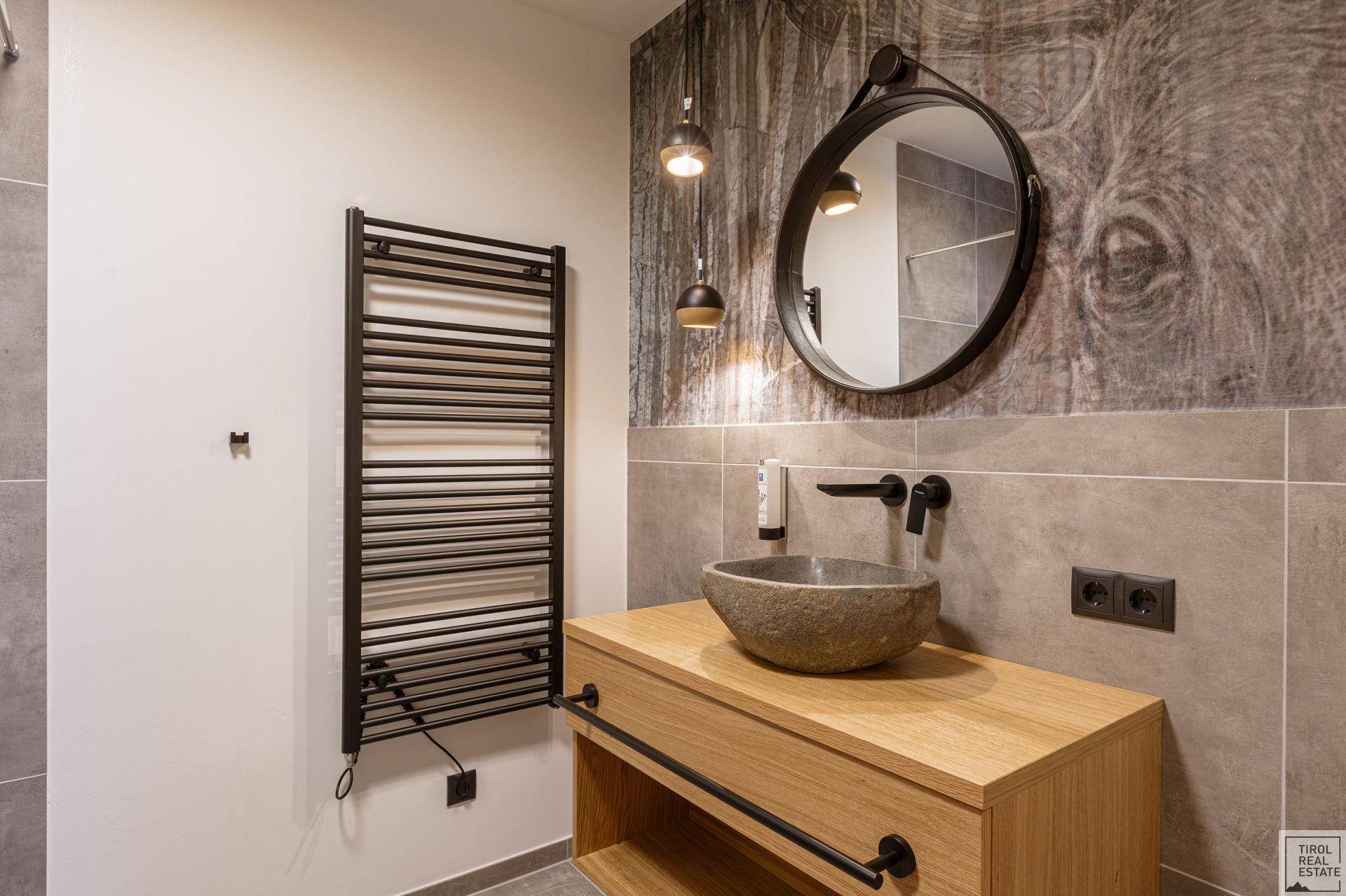 Modernes Badezimmer mit Steinswaschbecken, rundem Spiegel und beheiztem Handtuchhalter.
