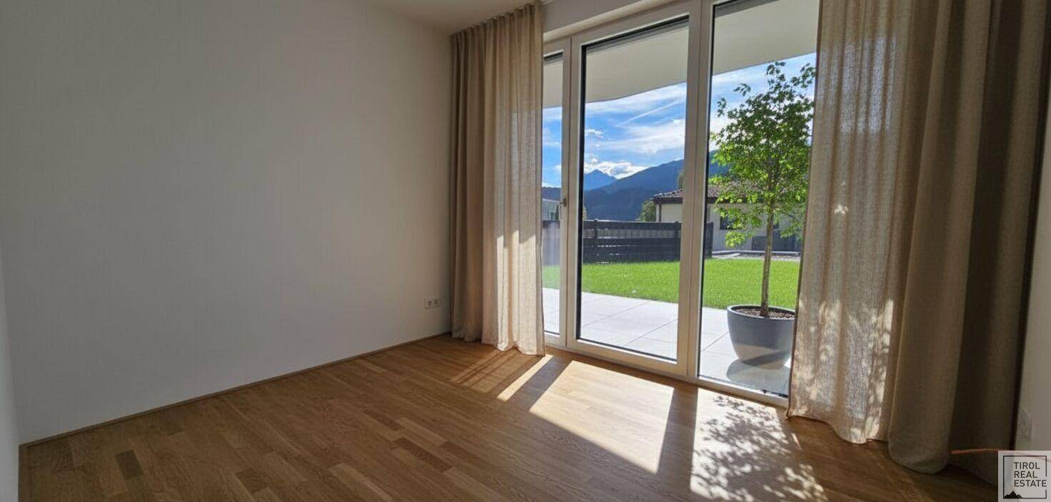 Leeres Zimmer mit Holzboden und großer Fensterfront zum Garten, bietet viel Tageslicht.