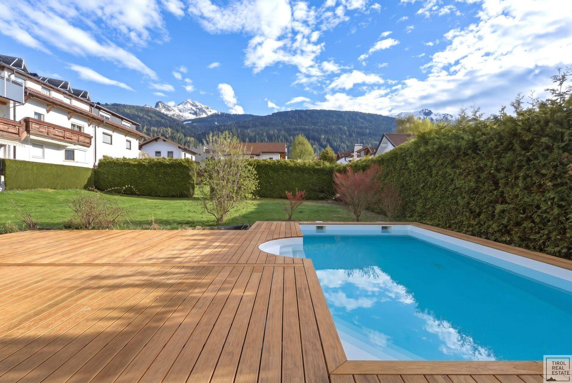 Sonniger Poolbereich mit Holzterrasse und Blick auf die Berge, umgeben von einer grünen Hecke.
