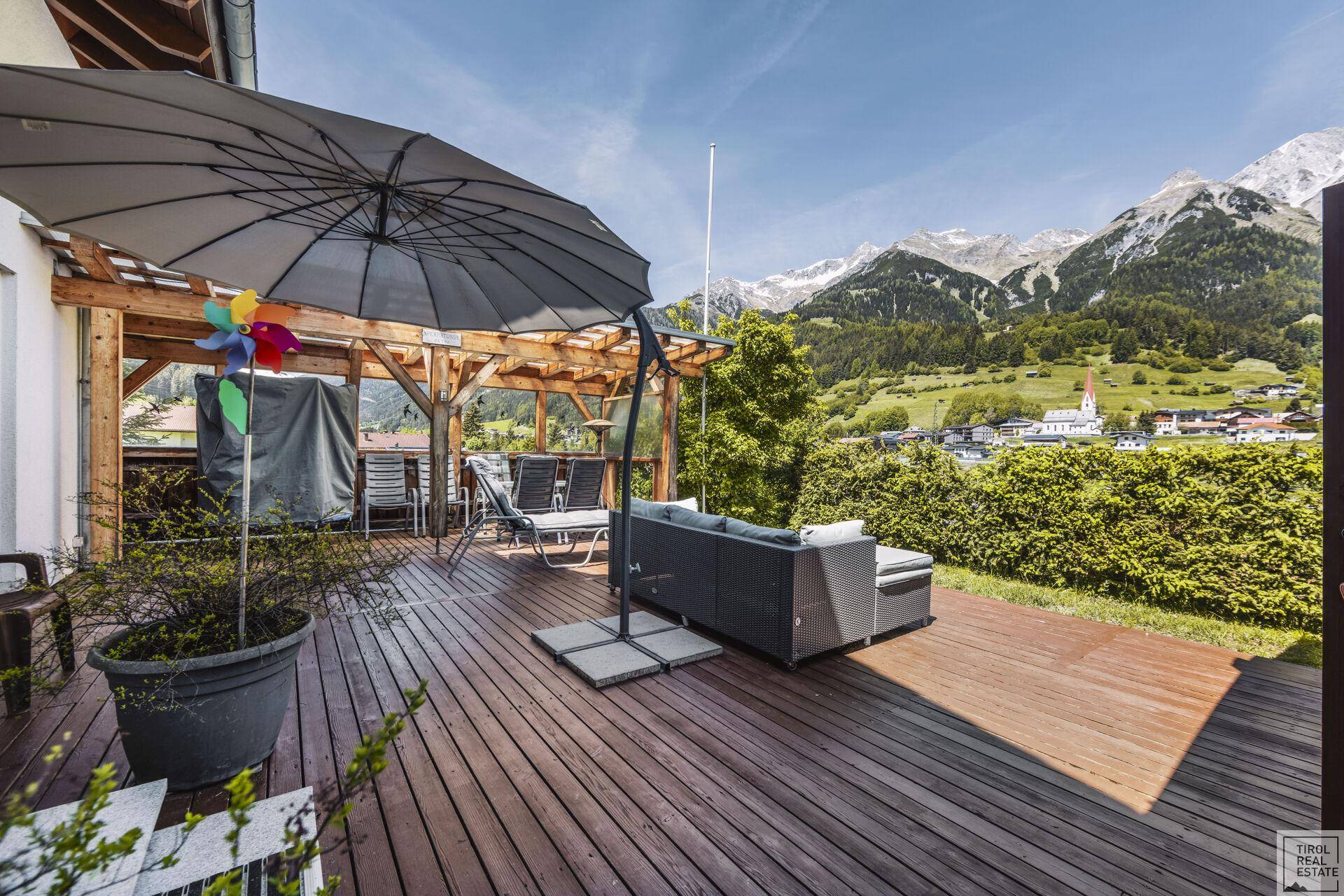 Große Holzterrasse mit Sonnenschirm und bequemen Lounge-Möbeln, umgeben von Natur.