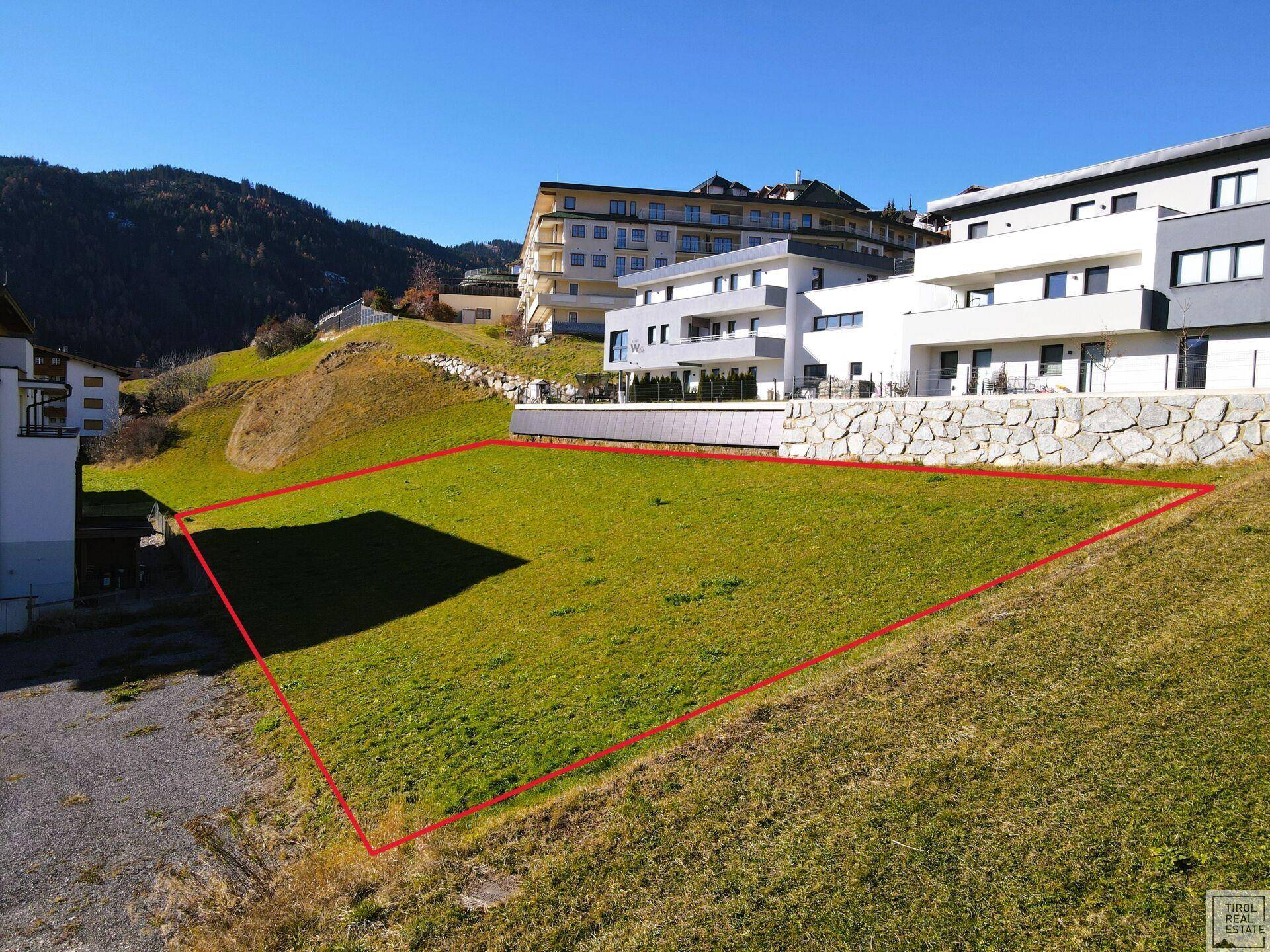 Blick auf ein sonniges Baugrundstück in grüner Umgebung mit modernen Wohngebäuden im Hintergrund.