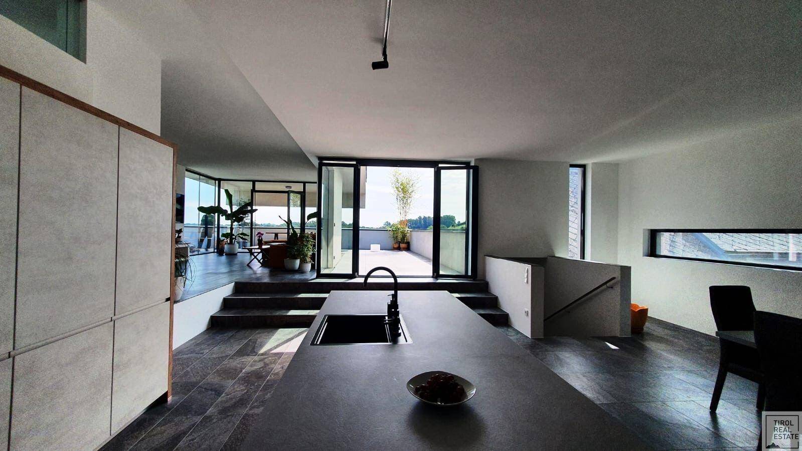 Offener Küchenbereich mit dunkler Arbeitsplatte, Blick auf den Wohnbereich und die Terrasse durch große Fenster.
