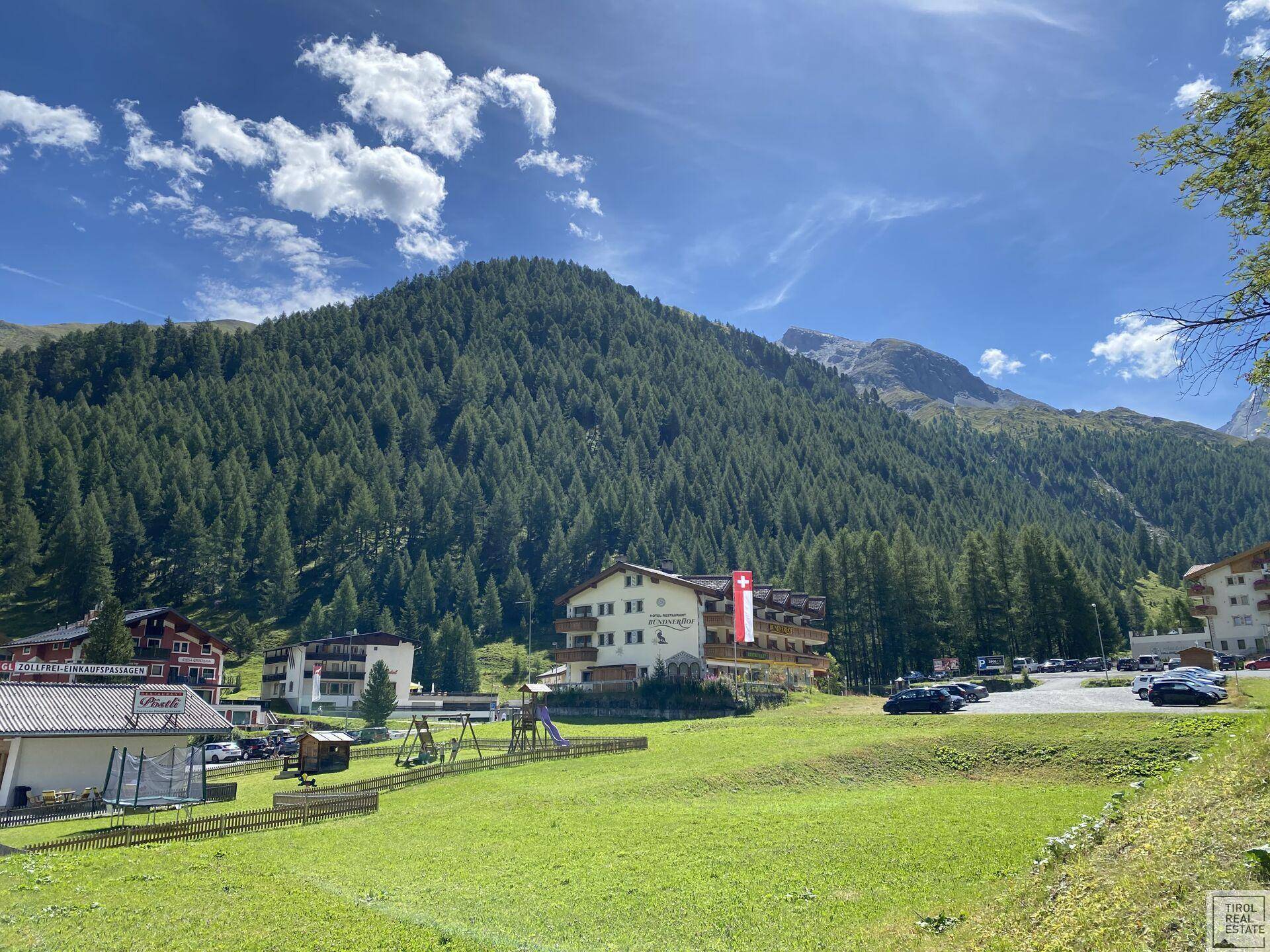 Hotelgebäude in einer grünen Berglandschaft mit Spielplatz und Parkplätzen unter blauem Himmel.