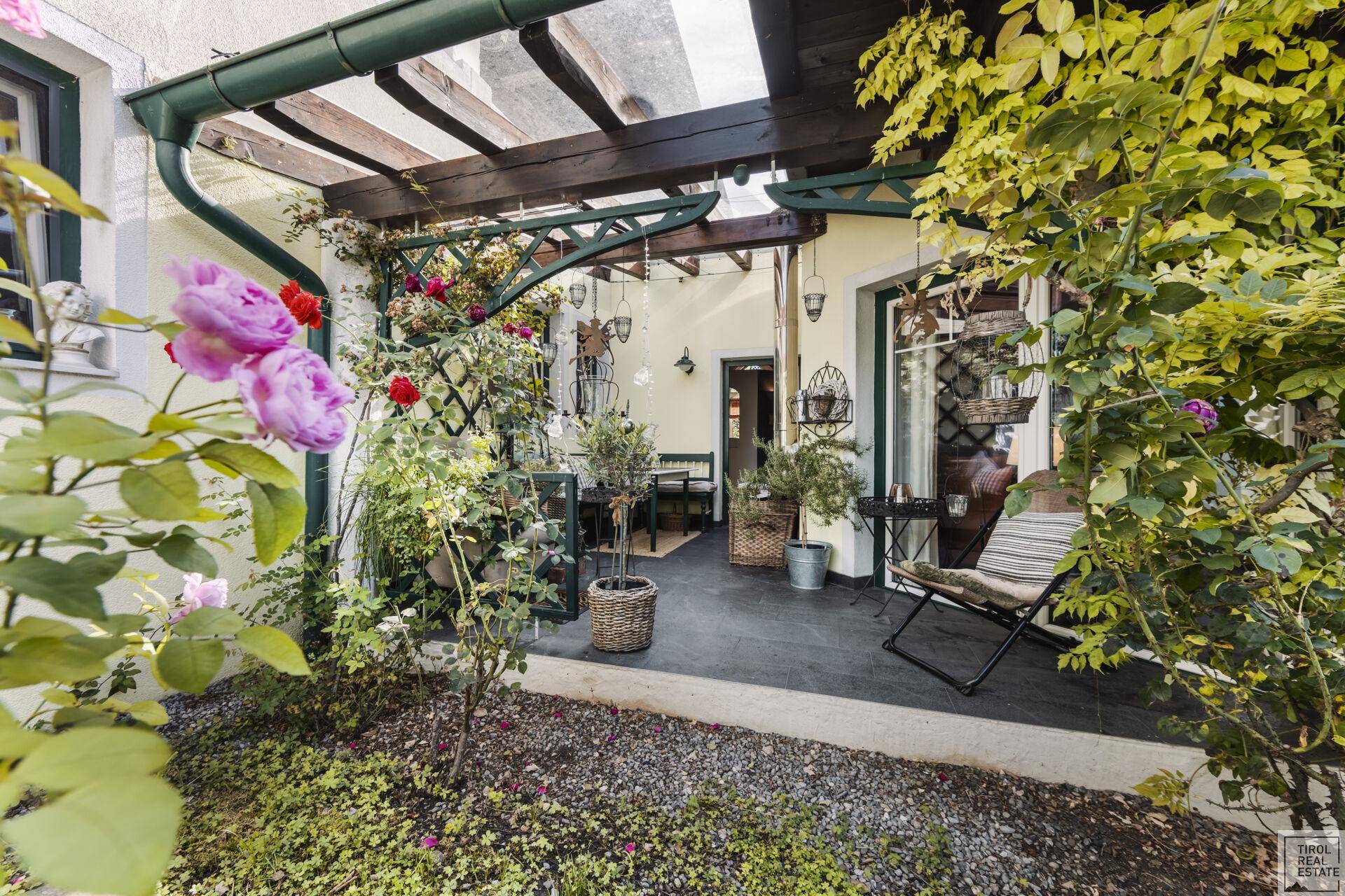 Charmante Terrasse mit Rosen im Vordergrund, überdachtem Sitzbereich und üppiger Bepflanzung.
