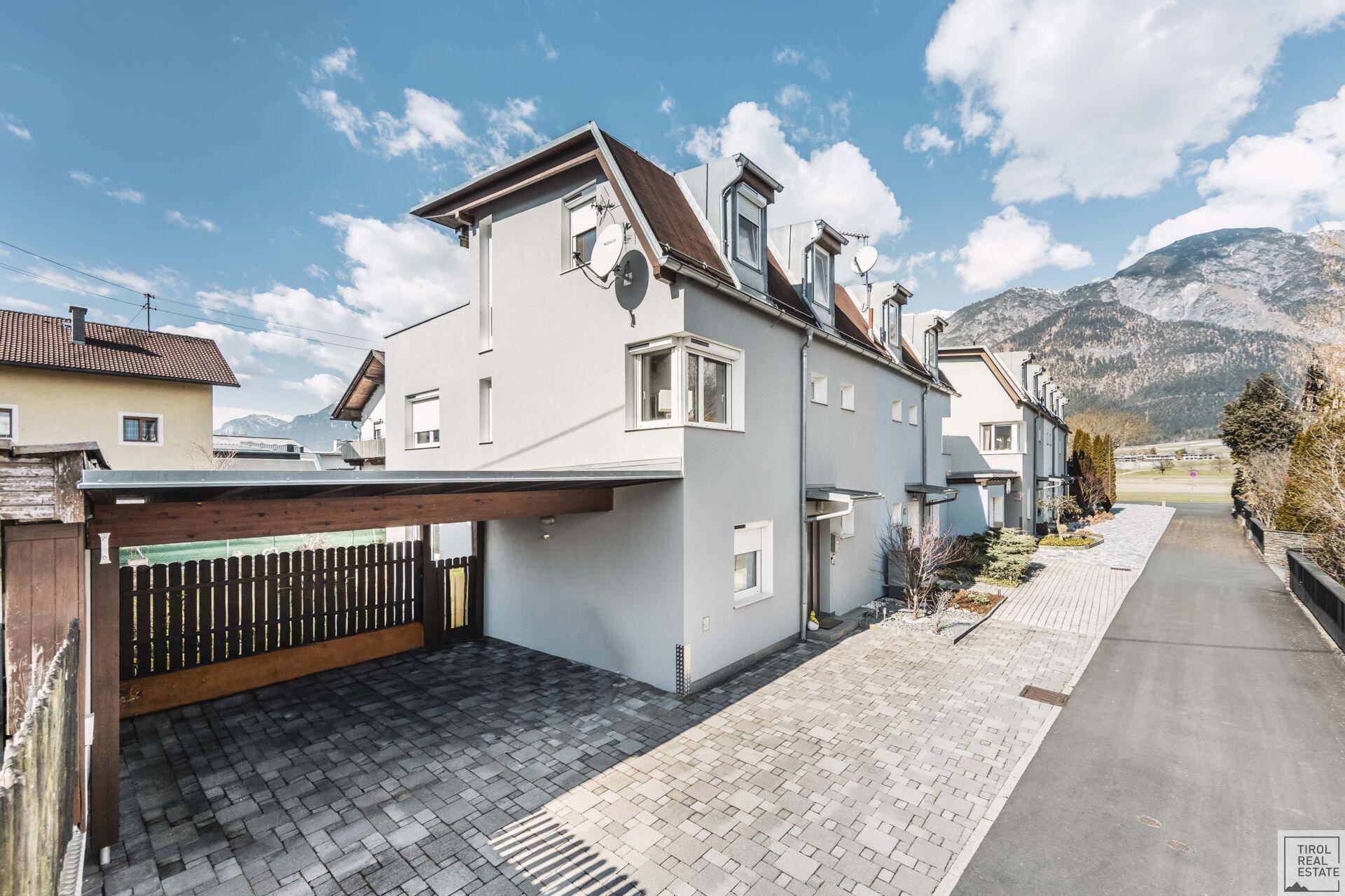 Moderne Reihenhausfassade mit Carport und gepflasterter Zufahrt vor einer beeindruckenden Bergkulisse.
