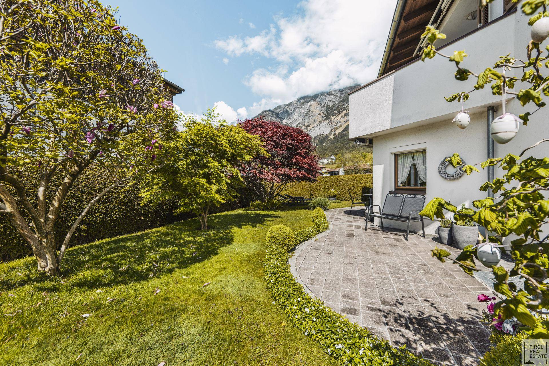 Gepflasterte Terrasse mit Sitzgelegenheiten, umgeben von einem grünen Garten und Bergblick.