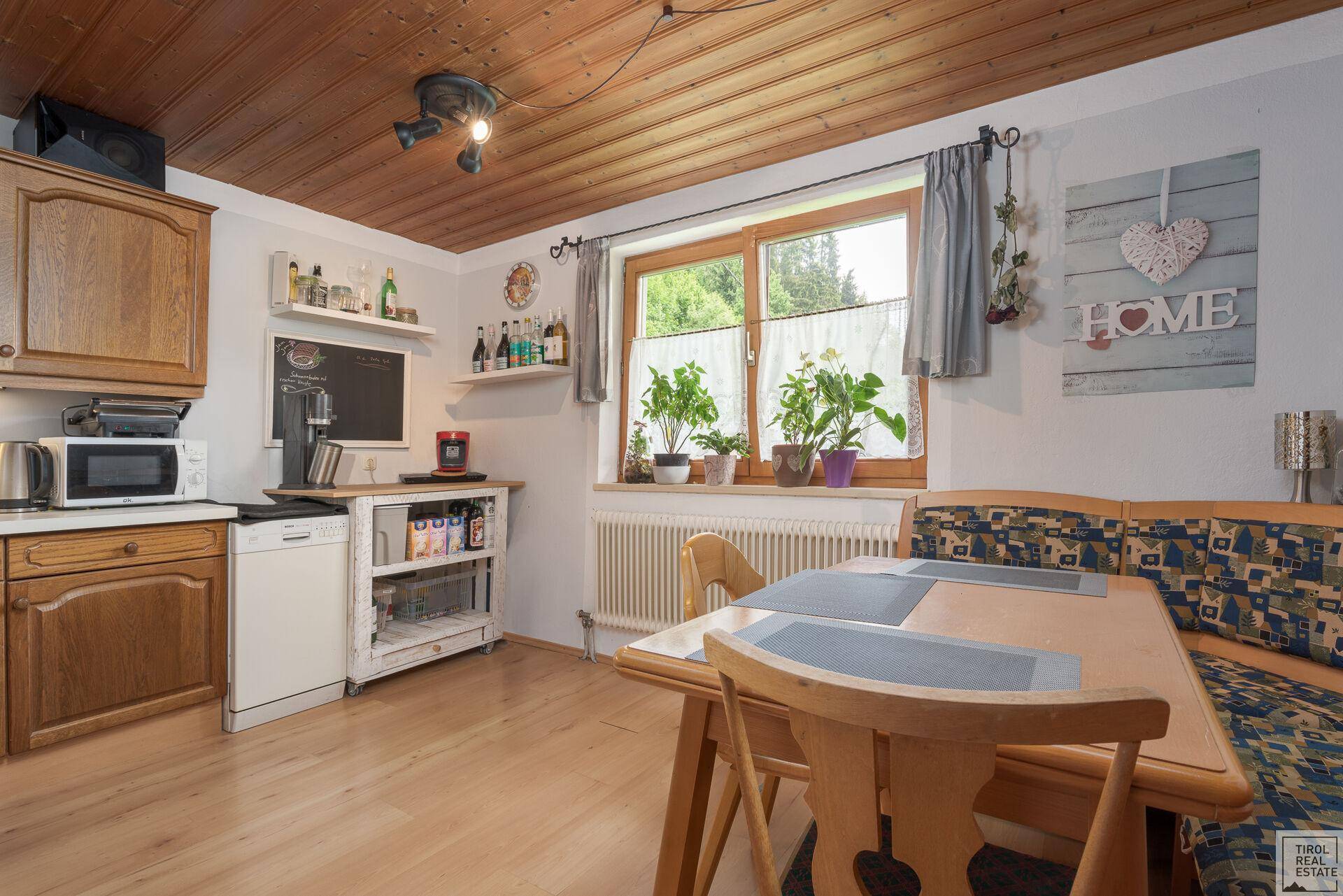 Rustikale Küche mit Holzeinbauten, Essbereich und Blick ins Grüne durch das Fenster.