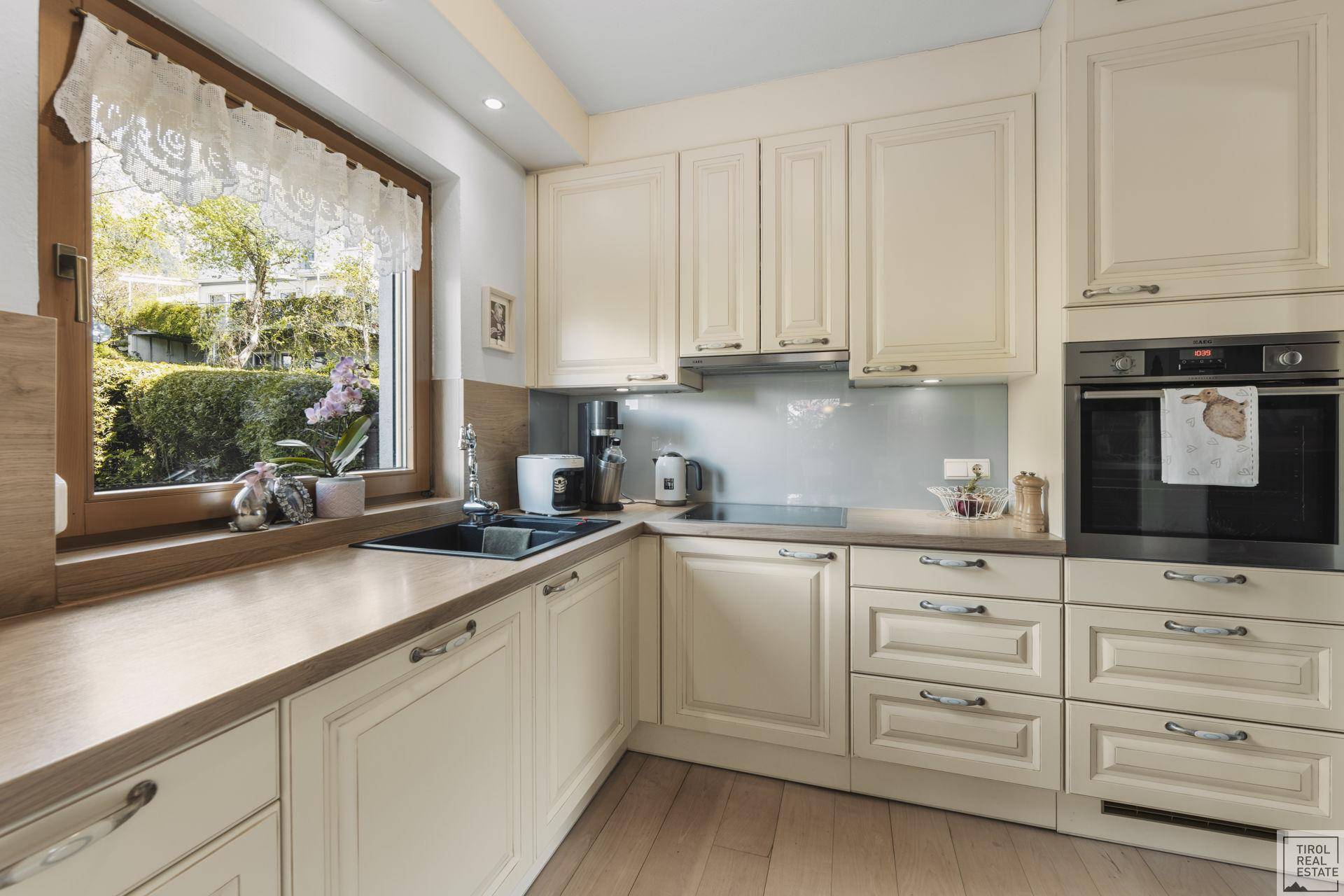 Helle Küche mit Fenster, Spüle, Kochfeld und Backofen, umgeben von cremefarbenen Schränken.