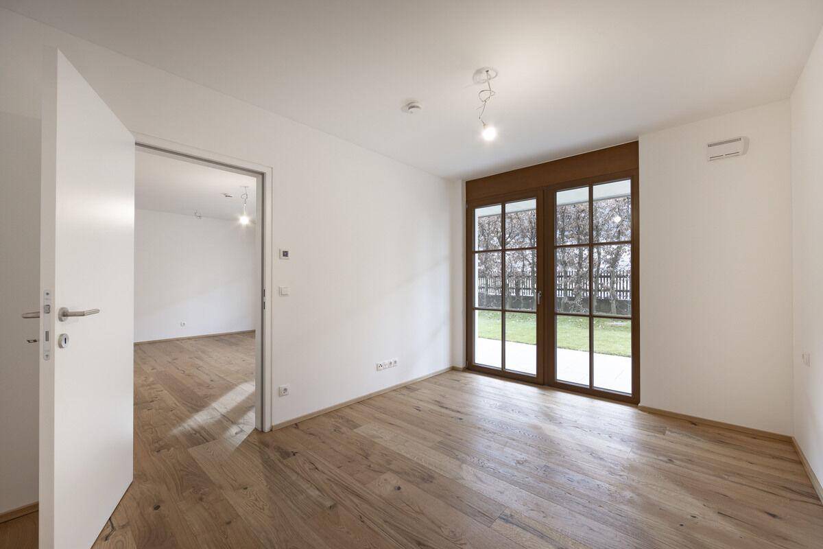 Lichtdurchfluteter Raum mit Holzboden und großen Fenstern, die den Blick auf den Außenbereich freigeben.