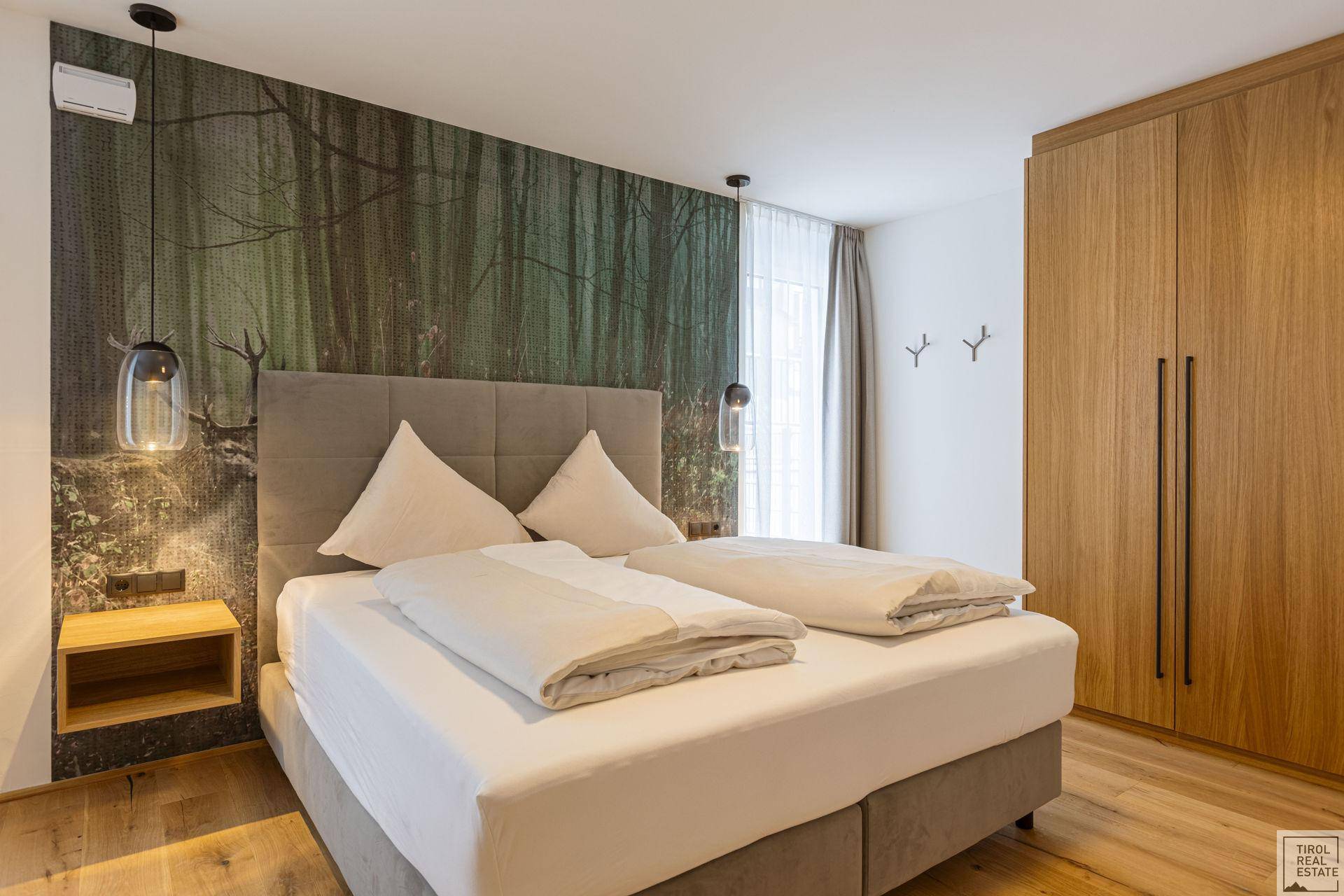 Schlafzimmer mit großem Doppelbett, Holzkleiderschrank und einer Waldtapete als Akzentwand.