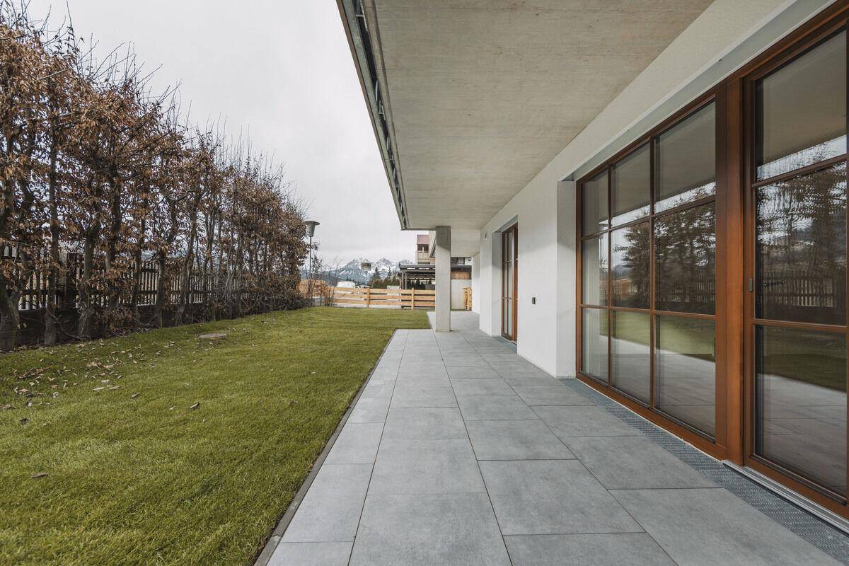 Große, geflieste Terrasse mit angrenzendem Rasenbereich und Hecke, ideal für Entspannung im Freien.