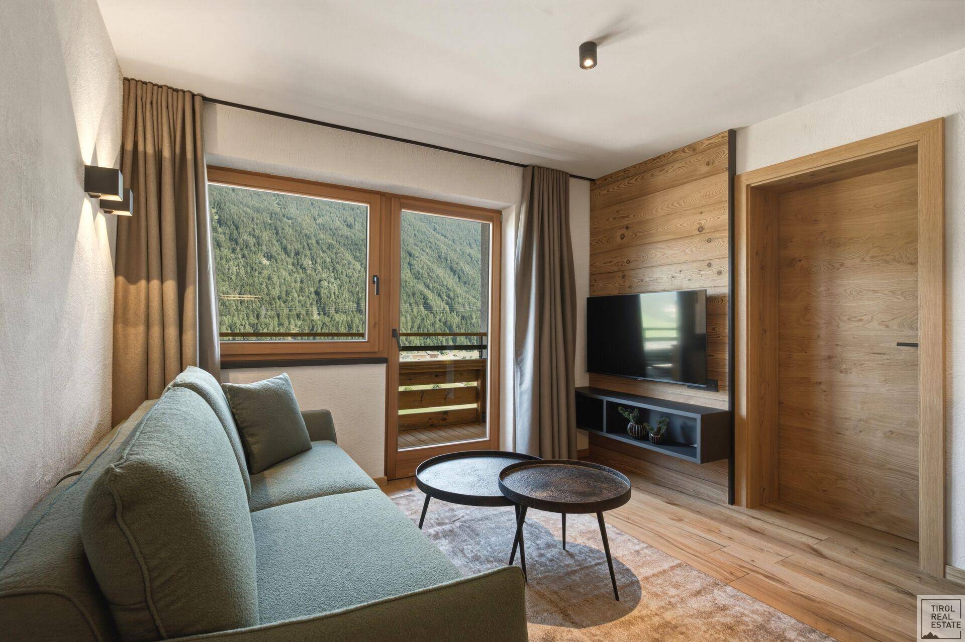 Gemütliches Wohnzimmer mit Sofa, Holzwand und Zugang zum Balkon mit Bergblick.