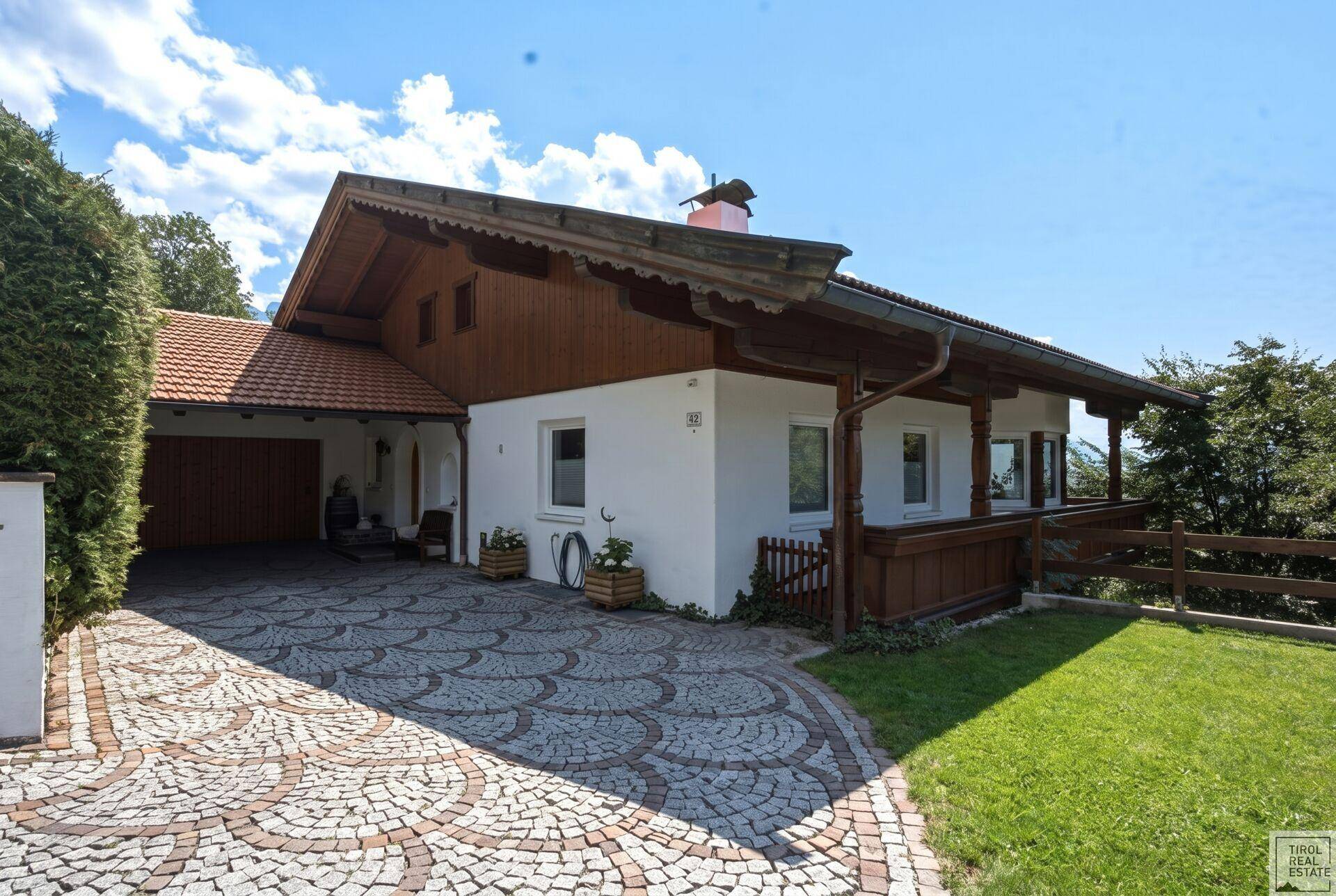 Traditionelles Haus mit weißer Fassade, Holzelementen und einem kunstvoll gepflasterten Vorplatz unter blauem Himmel.