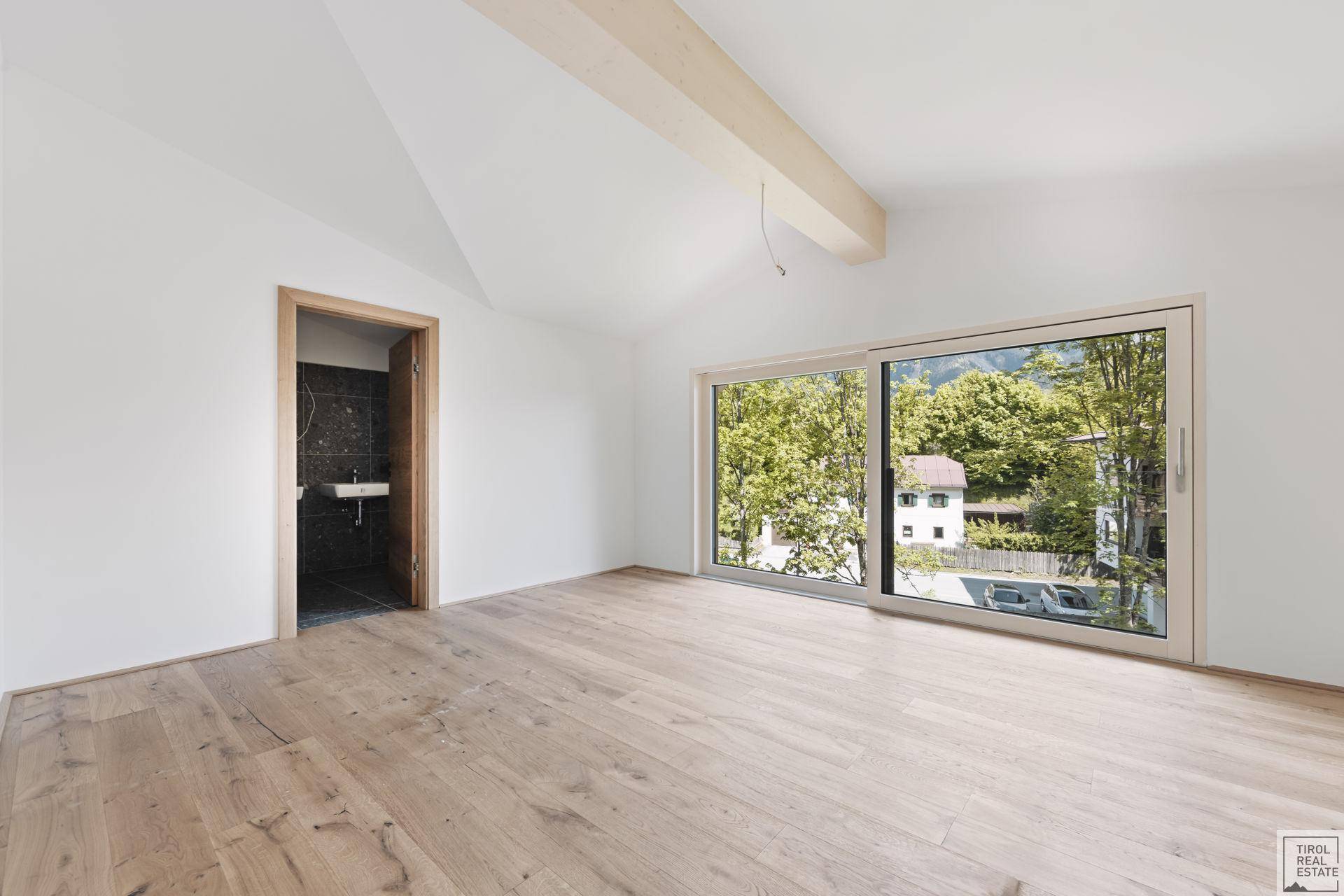 Helles Schlafzimmer mit Parkettboden, großem Fenster und Zugang zu einem Badezimmer.