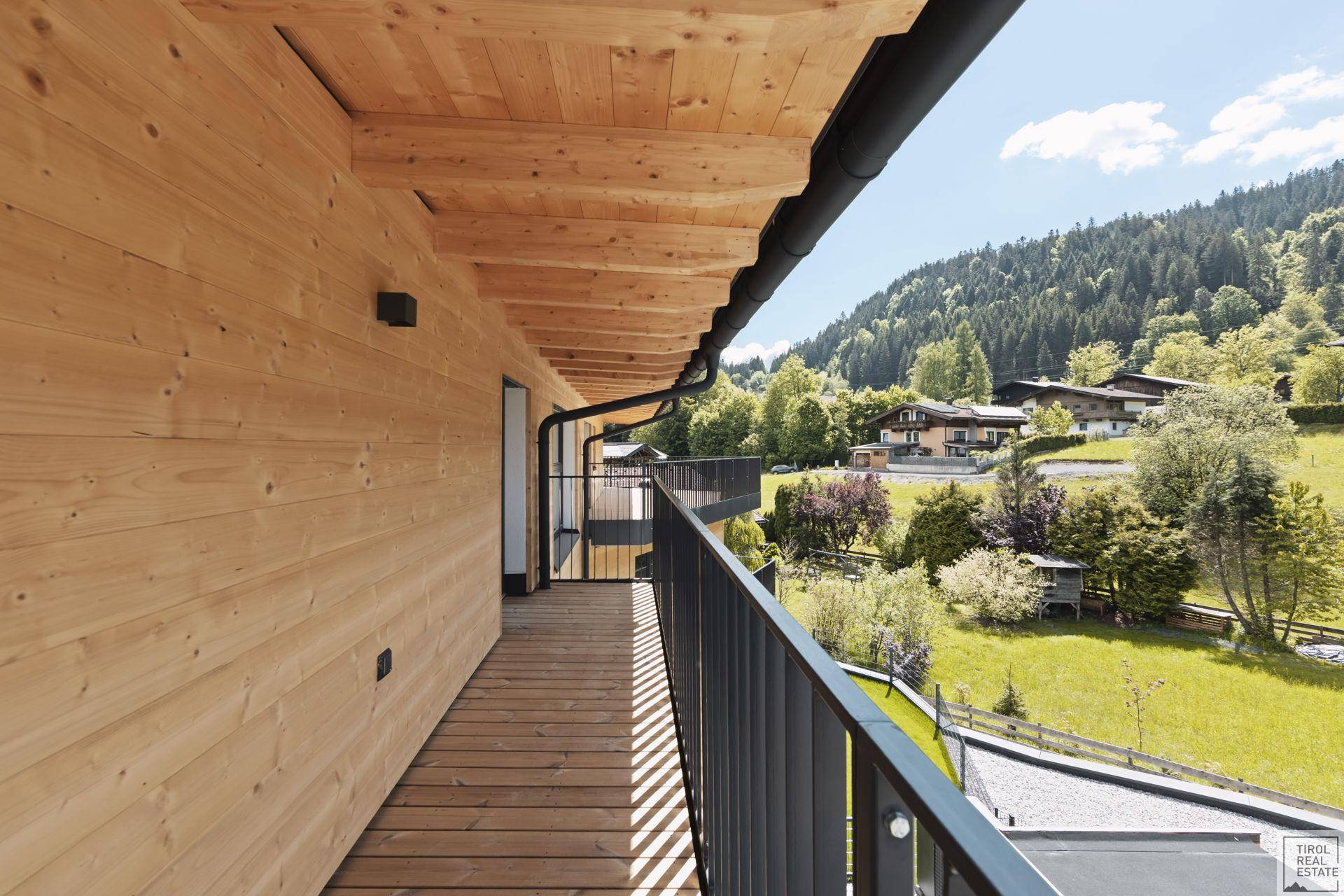 Schmaler Holzbalkon entlang der Fassade mit Blick auf grüne Wiesen und Häuser.