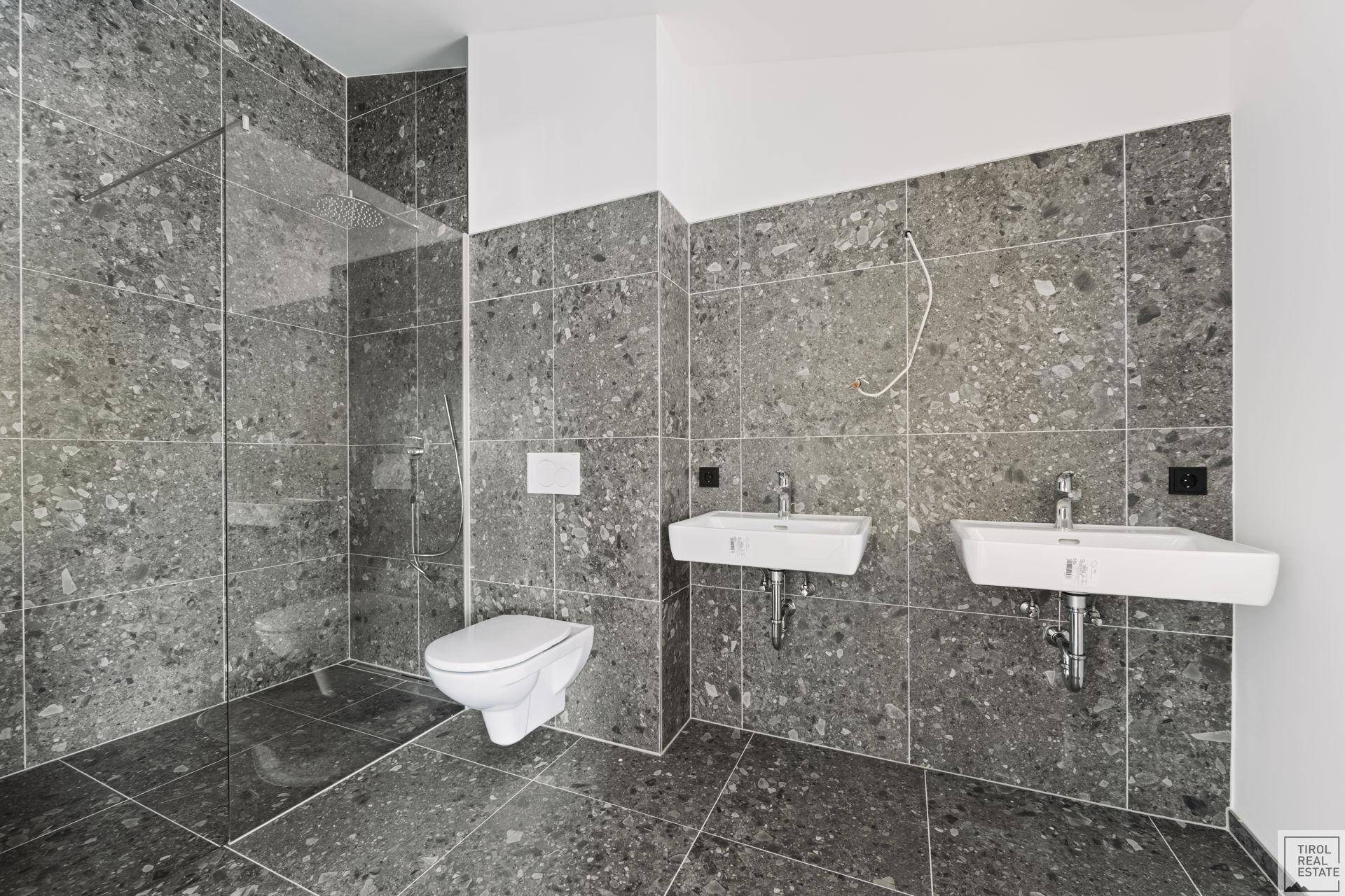 Modernes Badezimmer mit großformatigen grauen Fliesen, Dusche und zwei Waschbecken.
