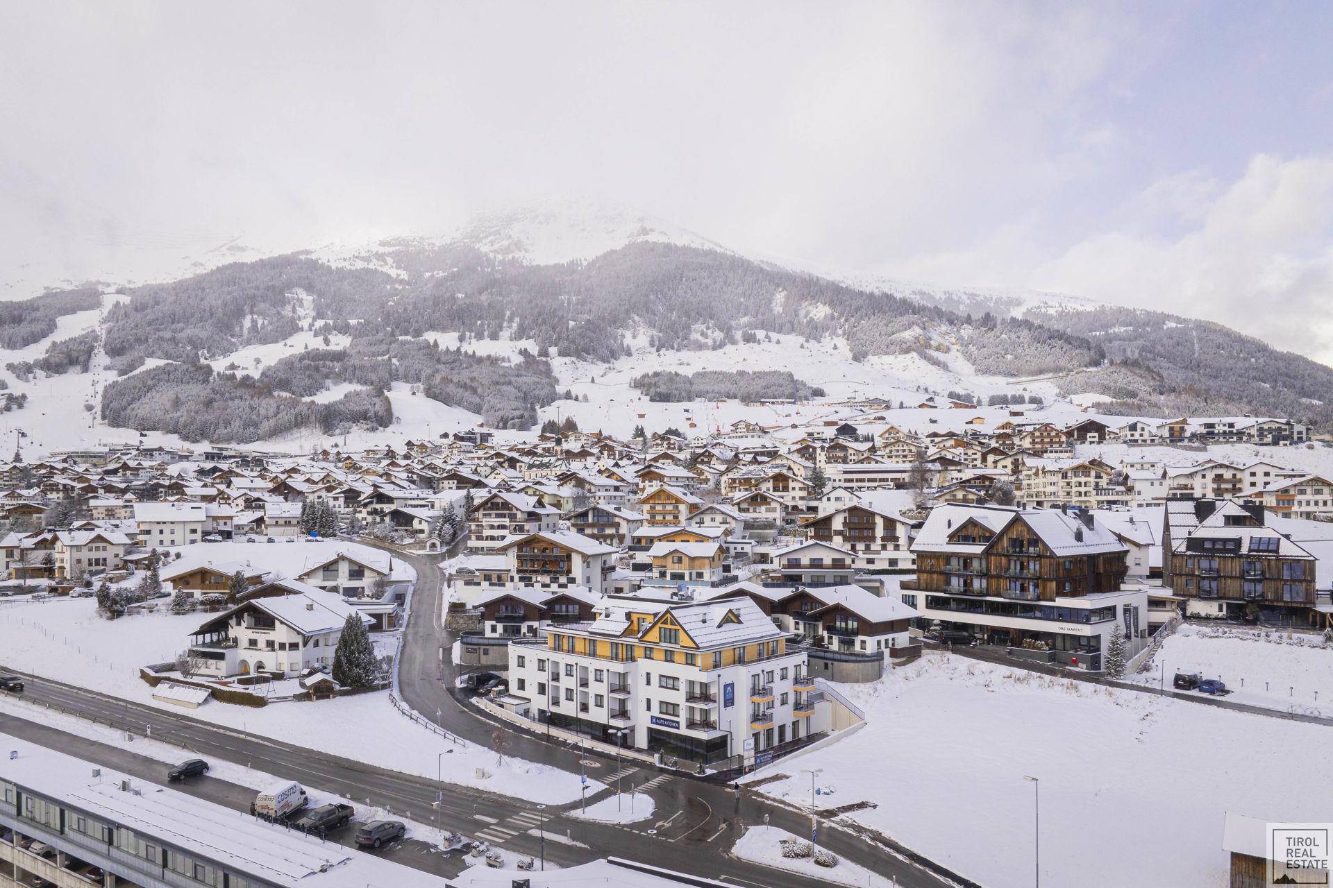 Weitläufiger Blick über ein schneebedecktes Dorf in den Alpen mit vielen Wohnhäusern und Bergen im Hintergrund.
