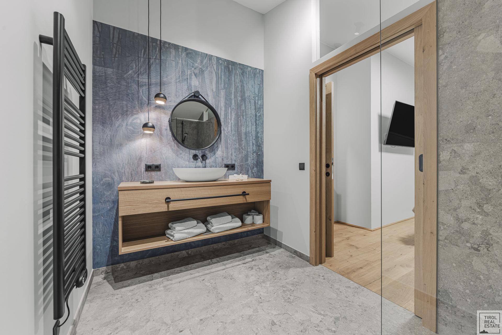 Modernes Badezimmer mit schwebendem Holzwaschtisch, rundem Spiegel und beheiztem Handtuchhalter.