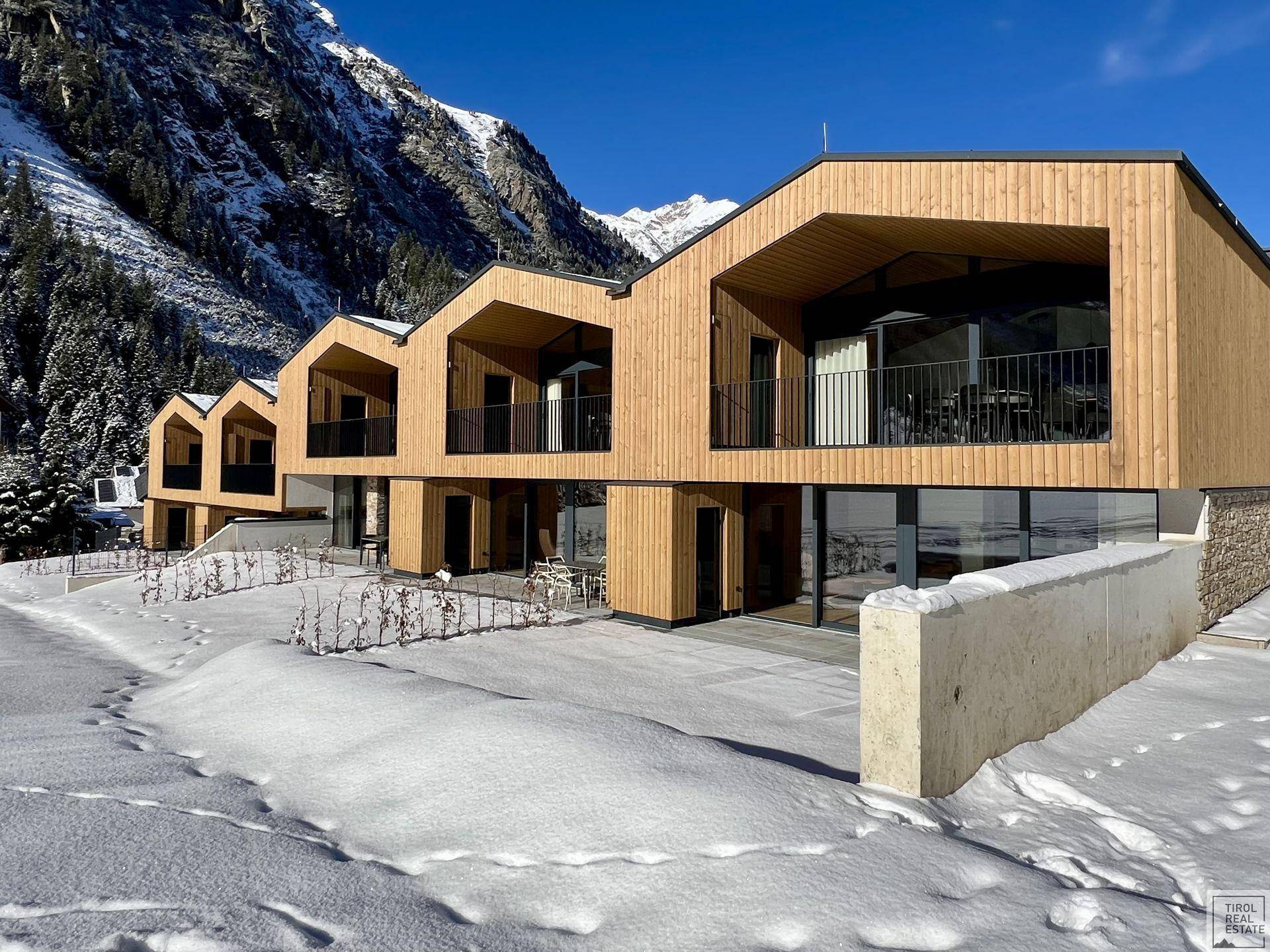 Reihe moderner Gebäude mit Holzverkleidung und großen Glasfronten in winterlicher Bergkulisse.