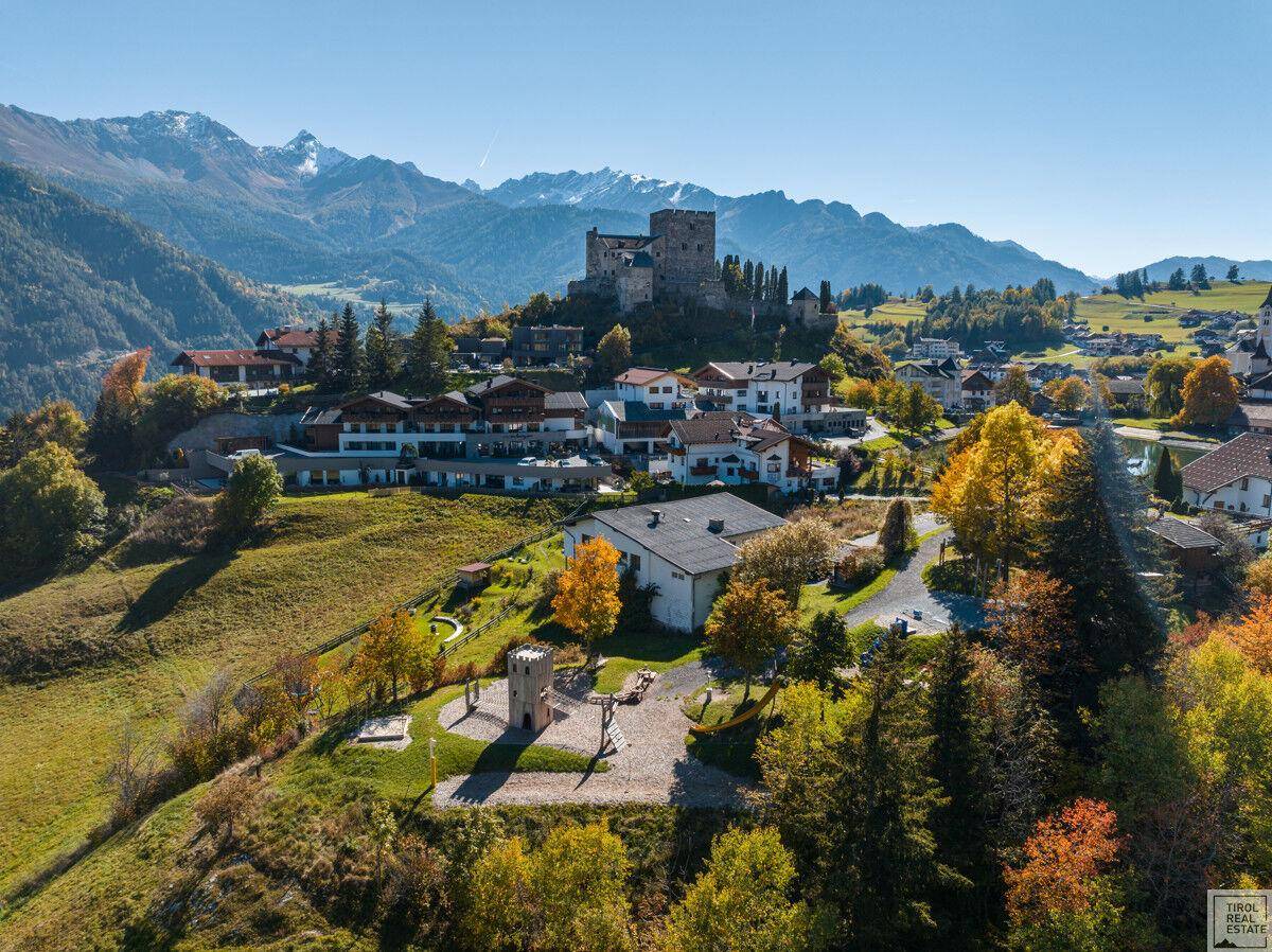 Idyllische Luftaufnahme eines Bergdorfes mit Burg, Spielplatz und beeindruckender Bergkulisse.