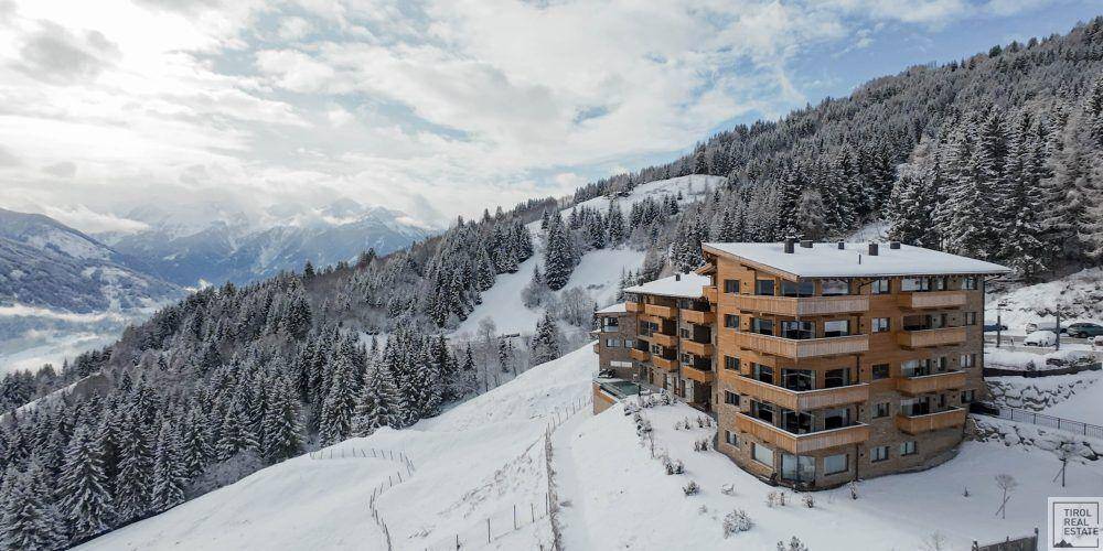 Gebäudekomplex auf einem verschneiten Berghang, umgeben von Tannenwäldern.