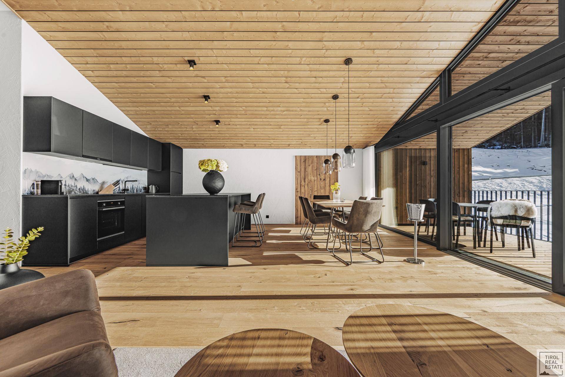 Offener Wohnbereich mit moderner schwarzer Küche, Holzbalkendecke und großen Fenstern mit Blick auf die verschneite Landschaft.
