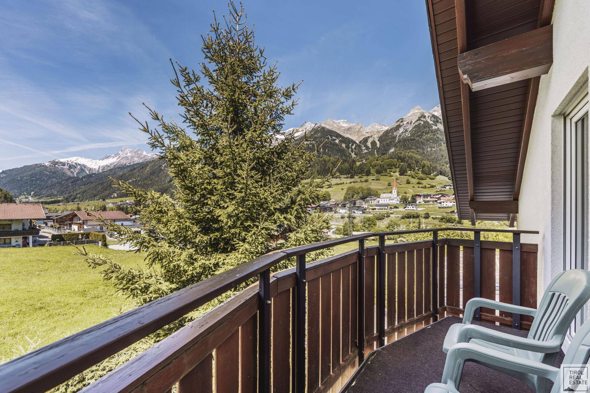 Balkon mit Holzgeländer und weitem Blick auf die umliegende Berglandschaft und das Dorf.