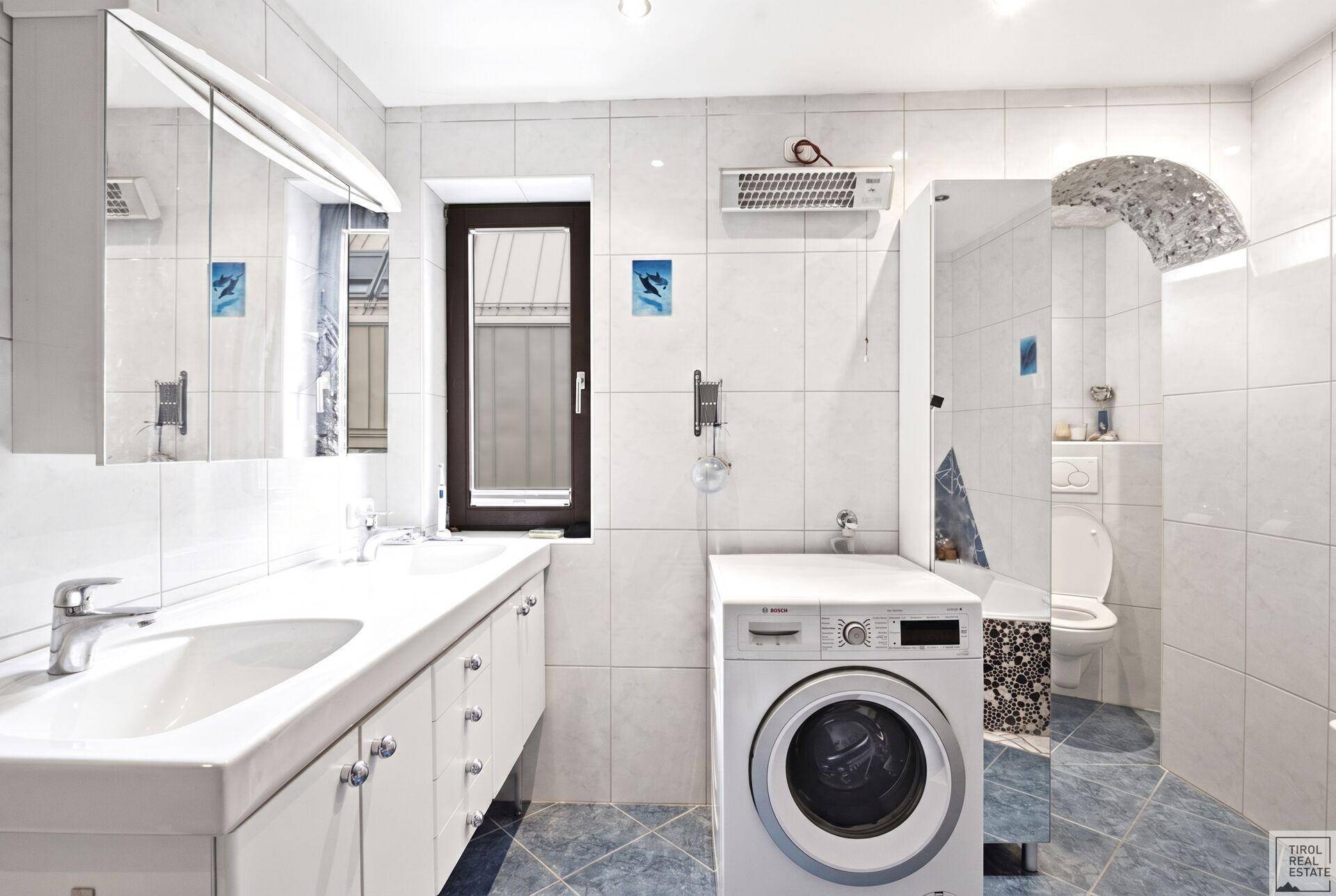 Gefliestes Badezimmer mit Doppelwaschbecken, Waschmaschine und Fenster.
