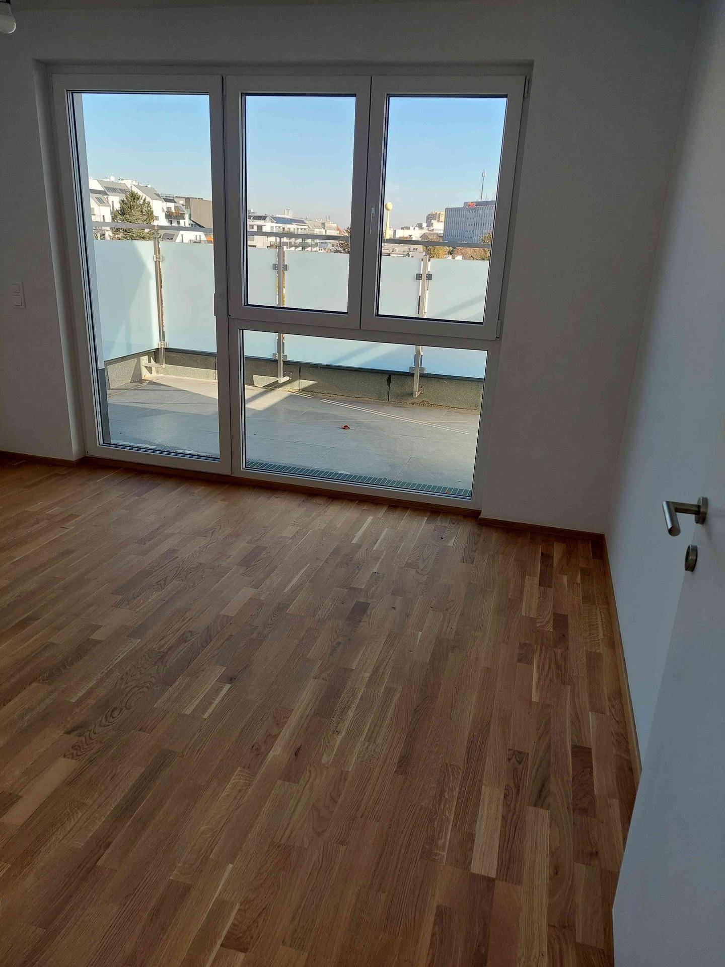 Helles Zimmer mit Holzboden und großen Fenstern, die den Blick auf die private Terrasse freigeben.