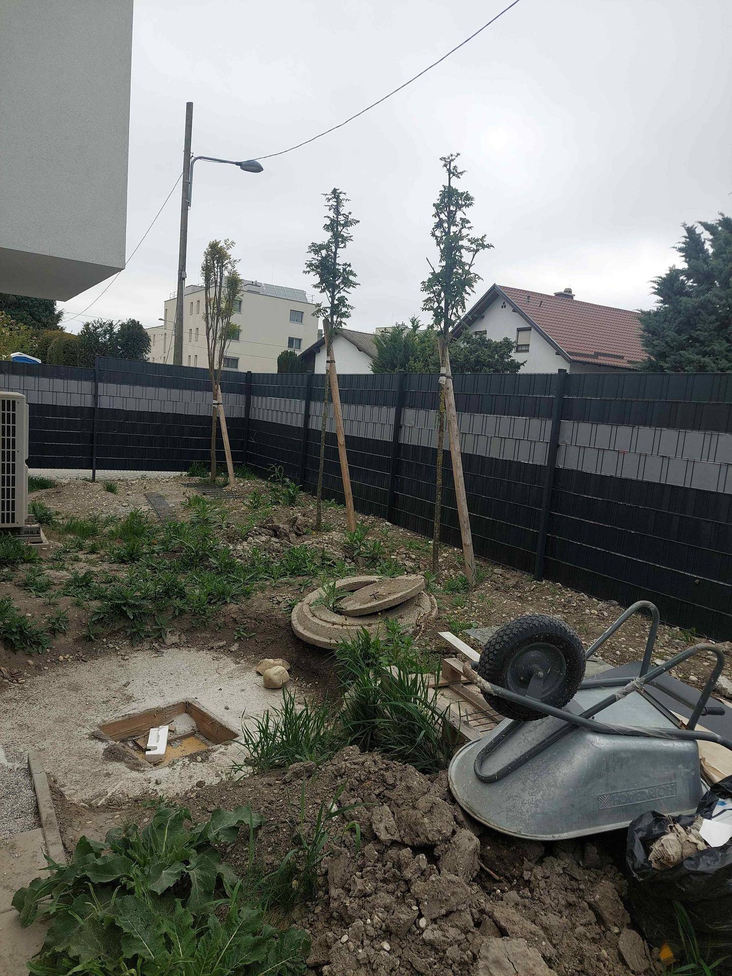 Gartenbereich im Bau mit jungen Bäumen, einer Schubkarre und einem blickdichten Zaun.