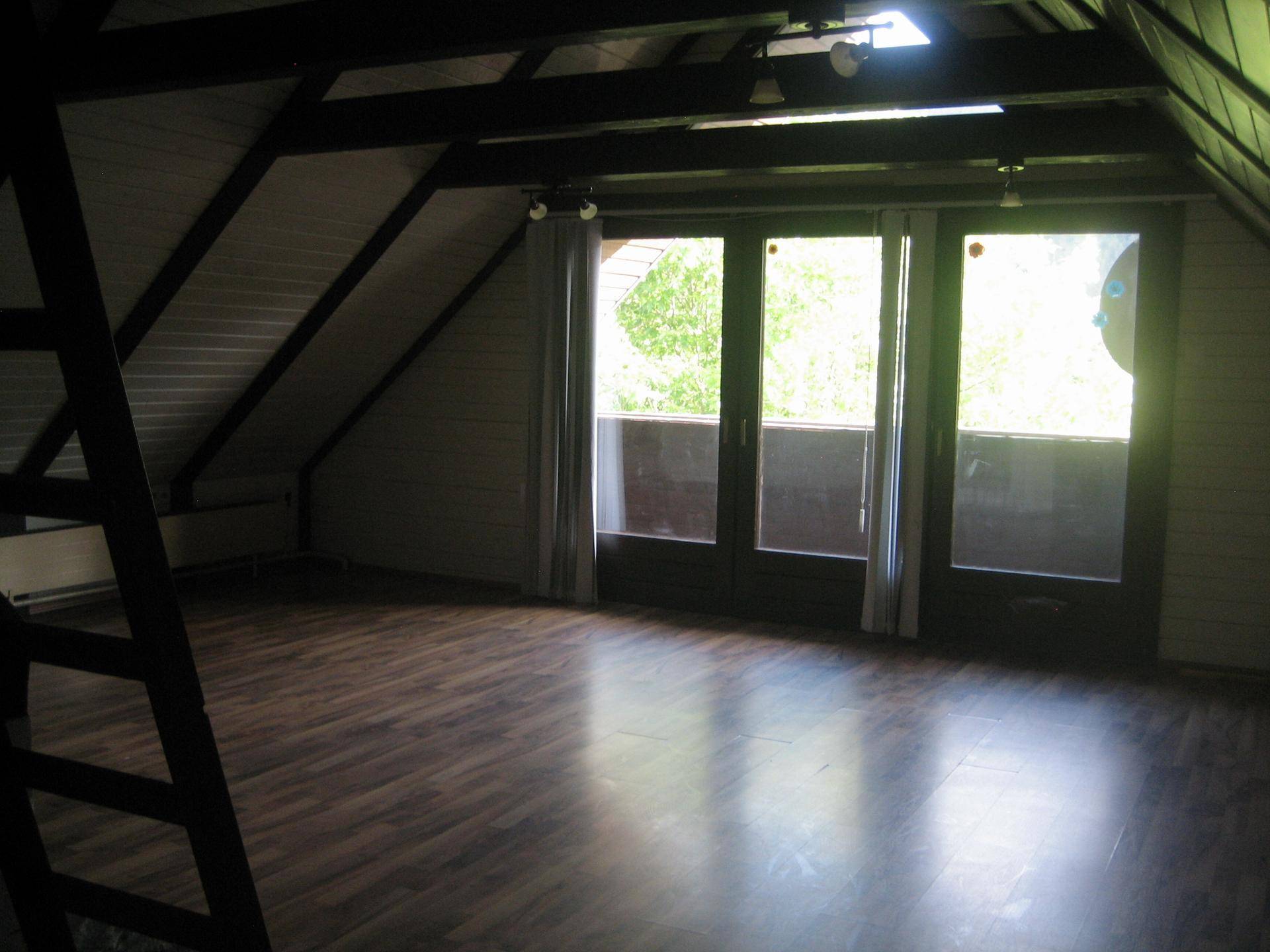 Geräumiger Dachgeschossraum mit Holzbalken, Laminatboden und großen Fenstern mit Blick ins Grüne.