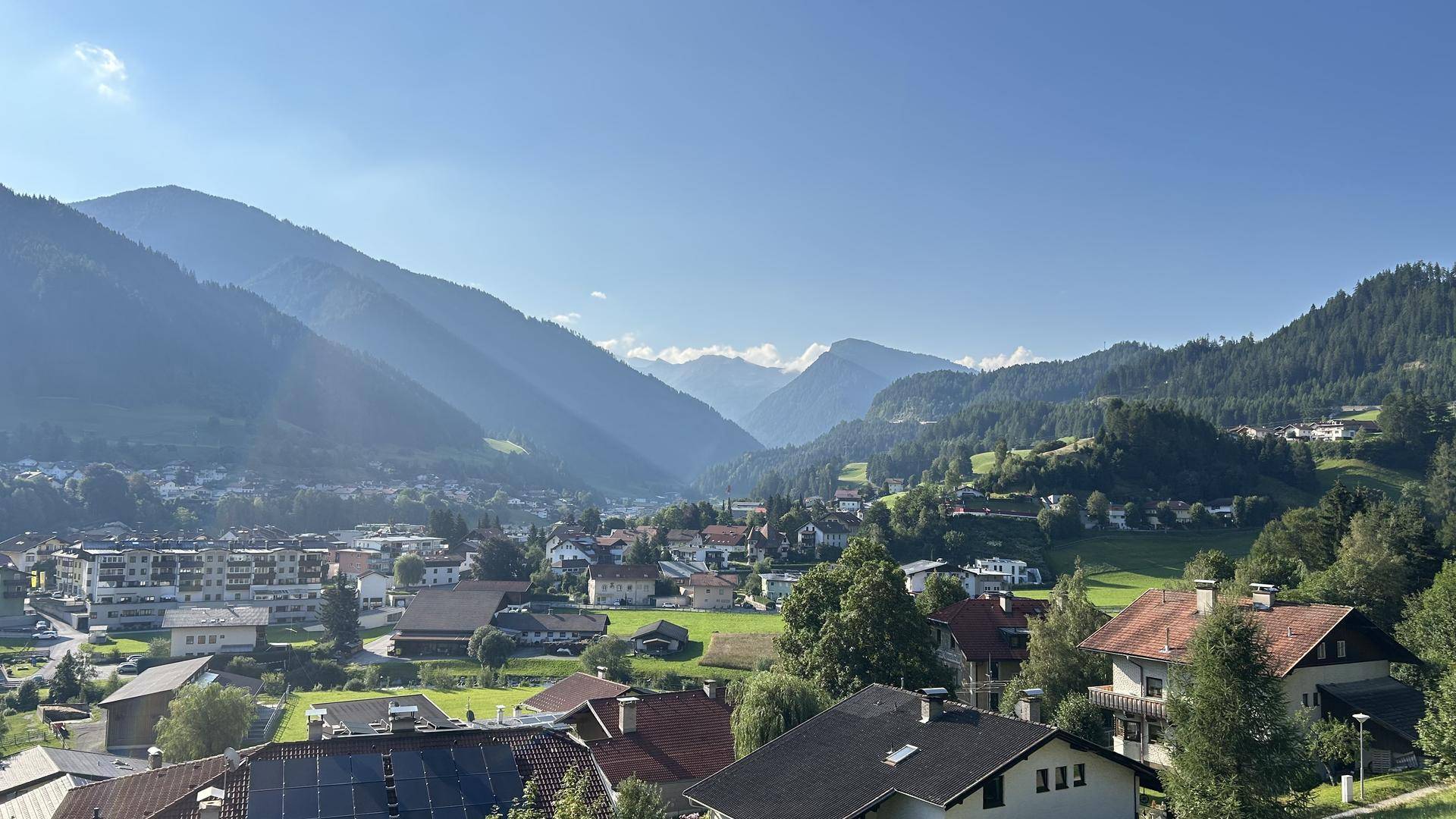Panoramablick über ein malerisches Tal mit Häusern und umliegenden Bergen unter blauem Himmel.