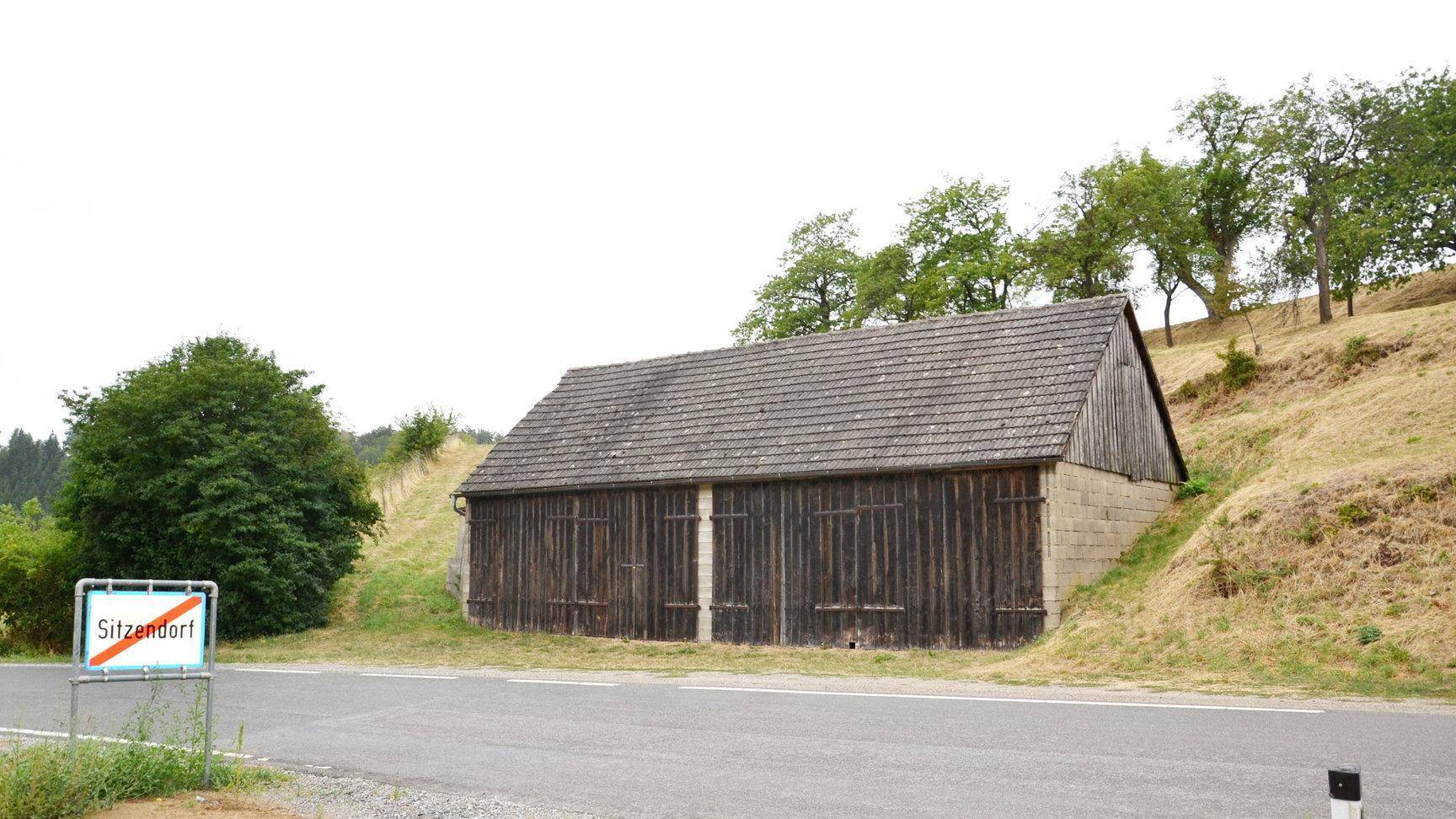 Altes Holzgebäude mit Ziegeldach an einer Landstraße, umgeben von Bäumen.