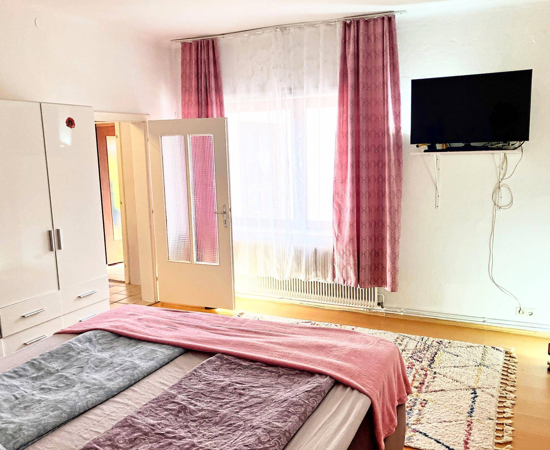 Schlafzimmer mit Doppelbett, weißem Kleiderschrank und rosa Vorhängen am Fenster.