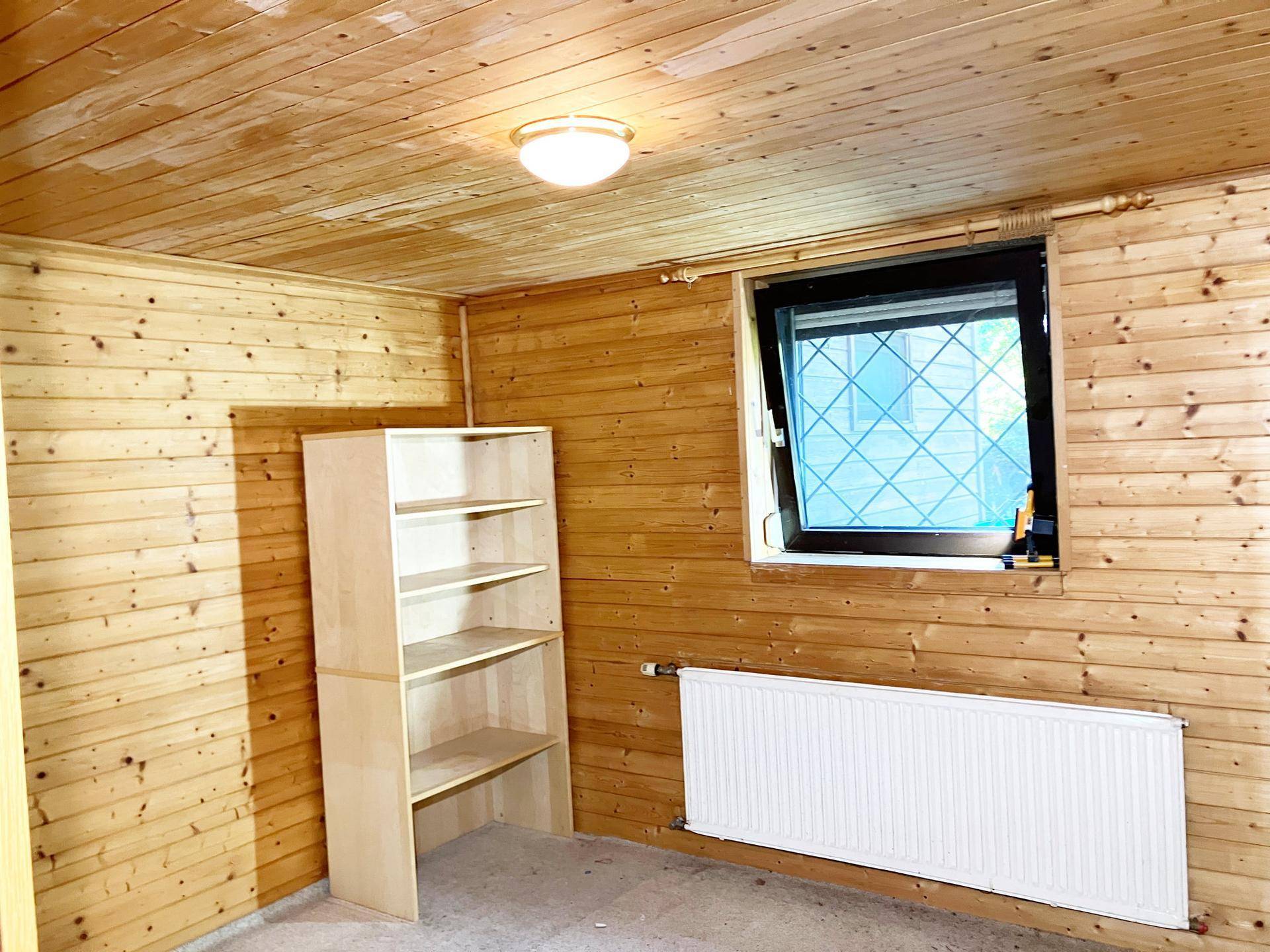 Helles Zimmer mit Holzverkleidung, einem Fenster, Heizkörper und einem einfachen Regal.
