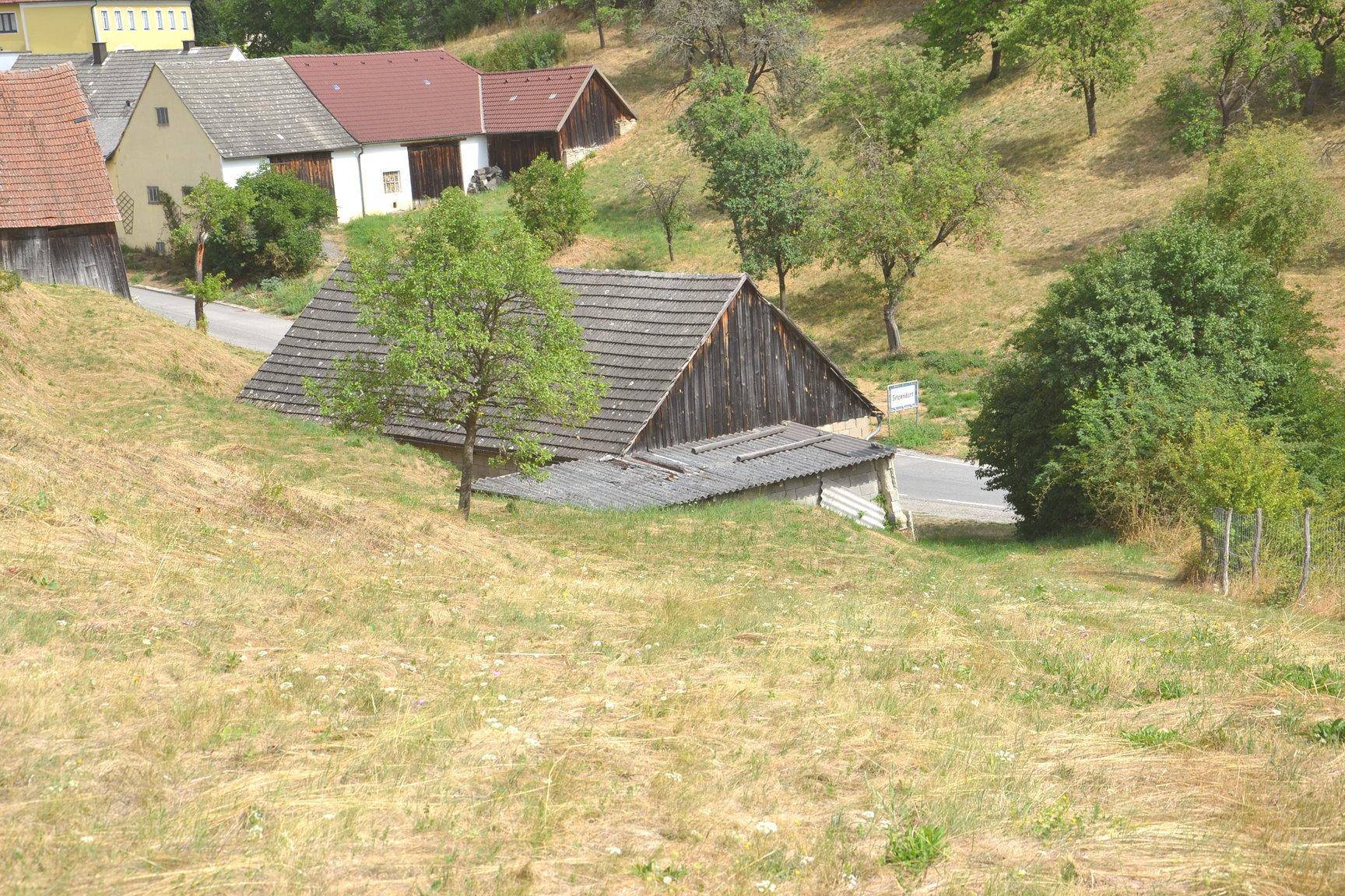 Blick über einen grasbewachsenen Hang auf ein altes Holzhaus und eine Straße im Tal.