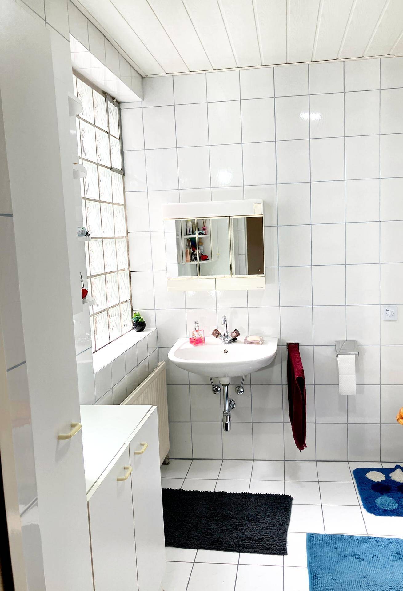 Helles Badezimmer mit weißen Fliesen, Einzelwaschbecken und einem Fenster aus Glasbausteinen.